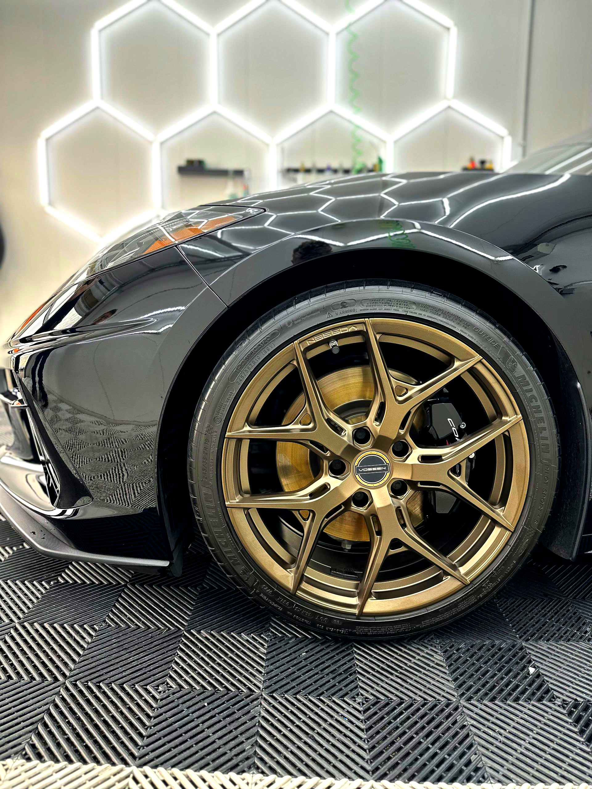 Close-up of a black sports car’s front wheel with gold rim on a patterned showroom floor
