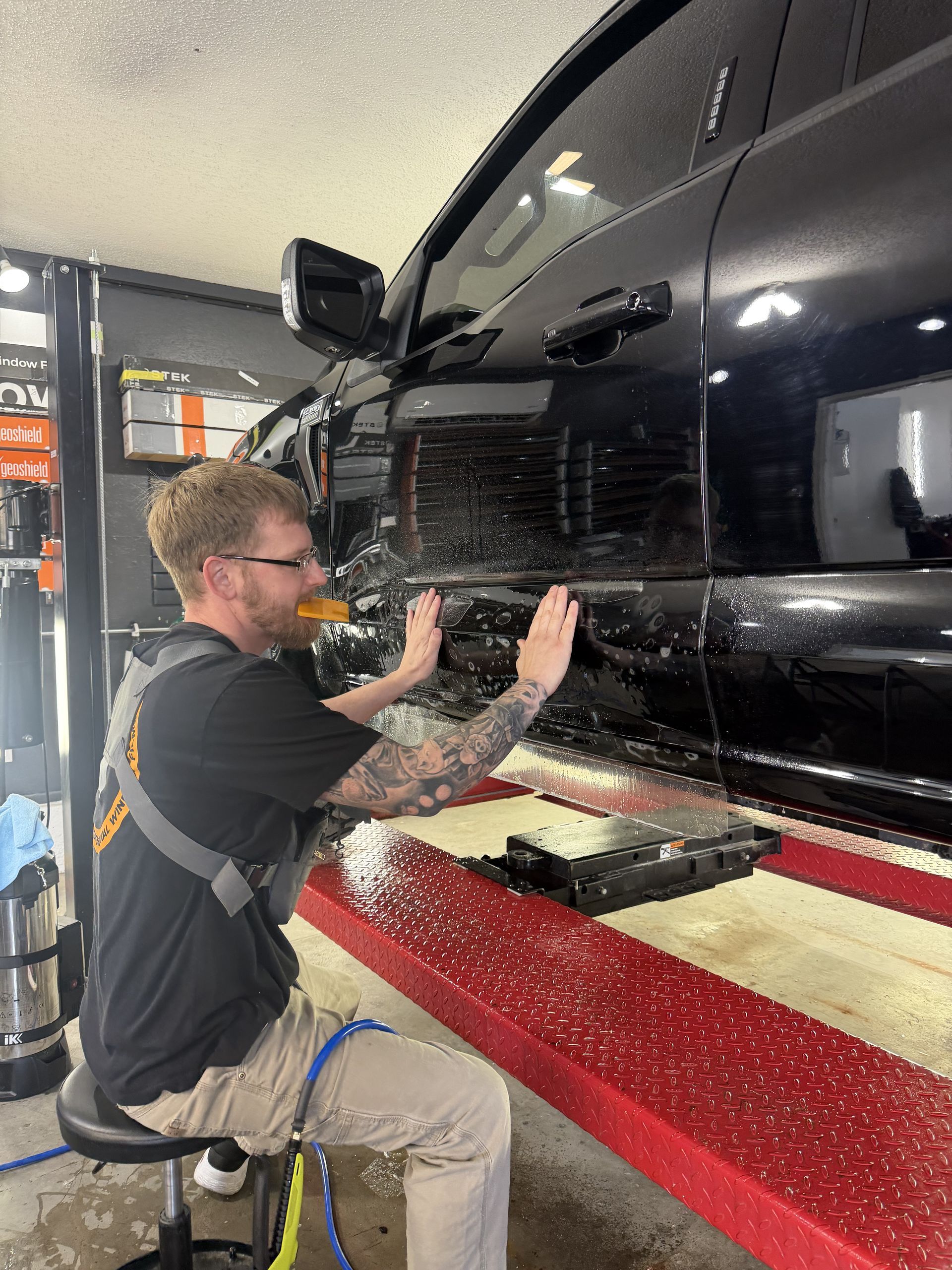 Mechanic polishing a black car in an auto shop with a red lift and tools nearby