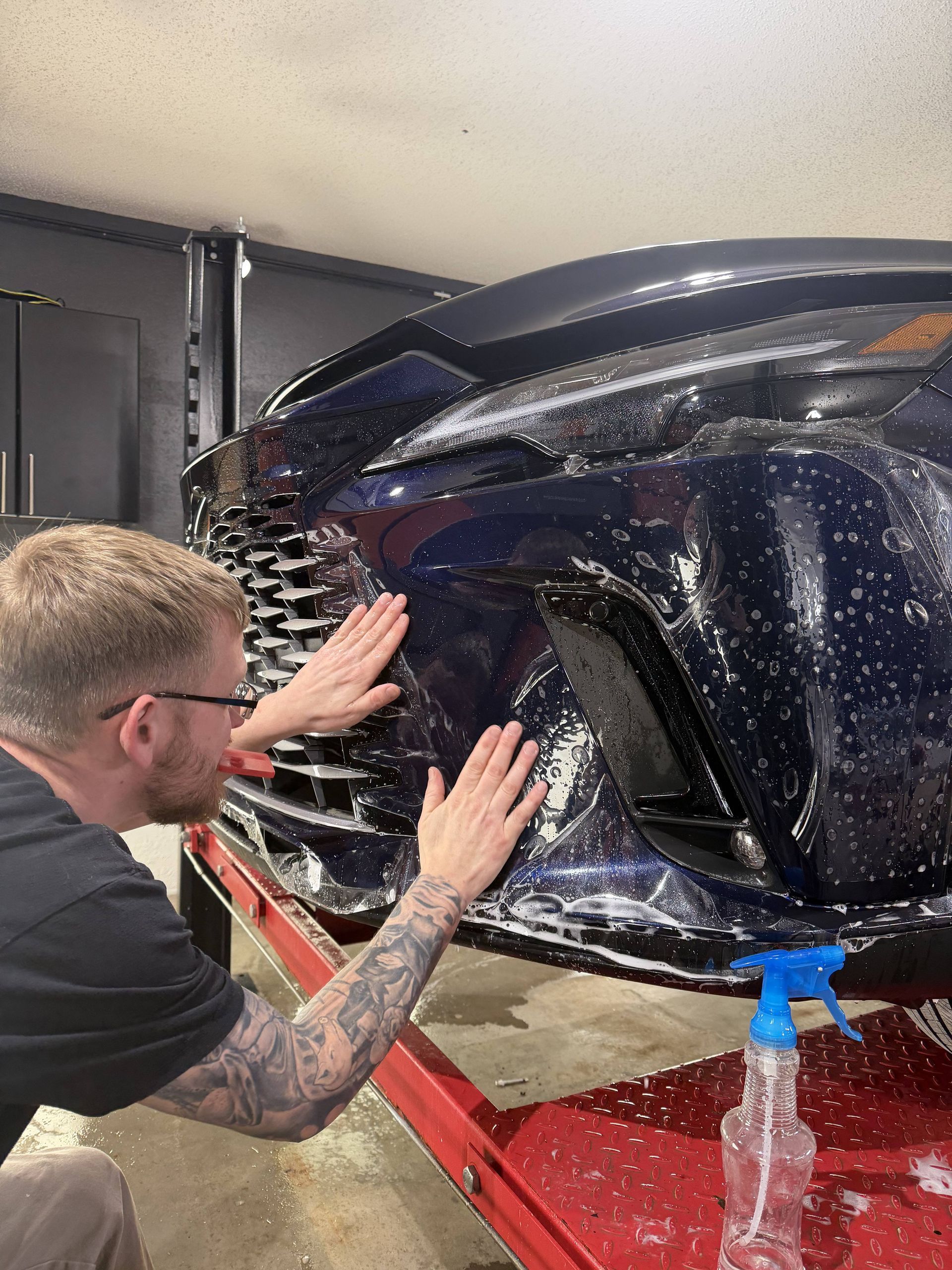 Man polishing a black car bumper in a garage with a spray bottle on the red lift.