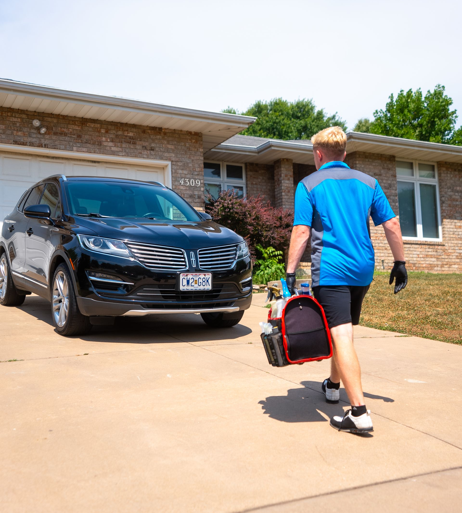 Person in blue shirt walks towards house carrying equipment bag, black car parked in driveway.