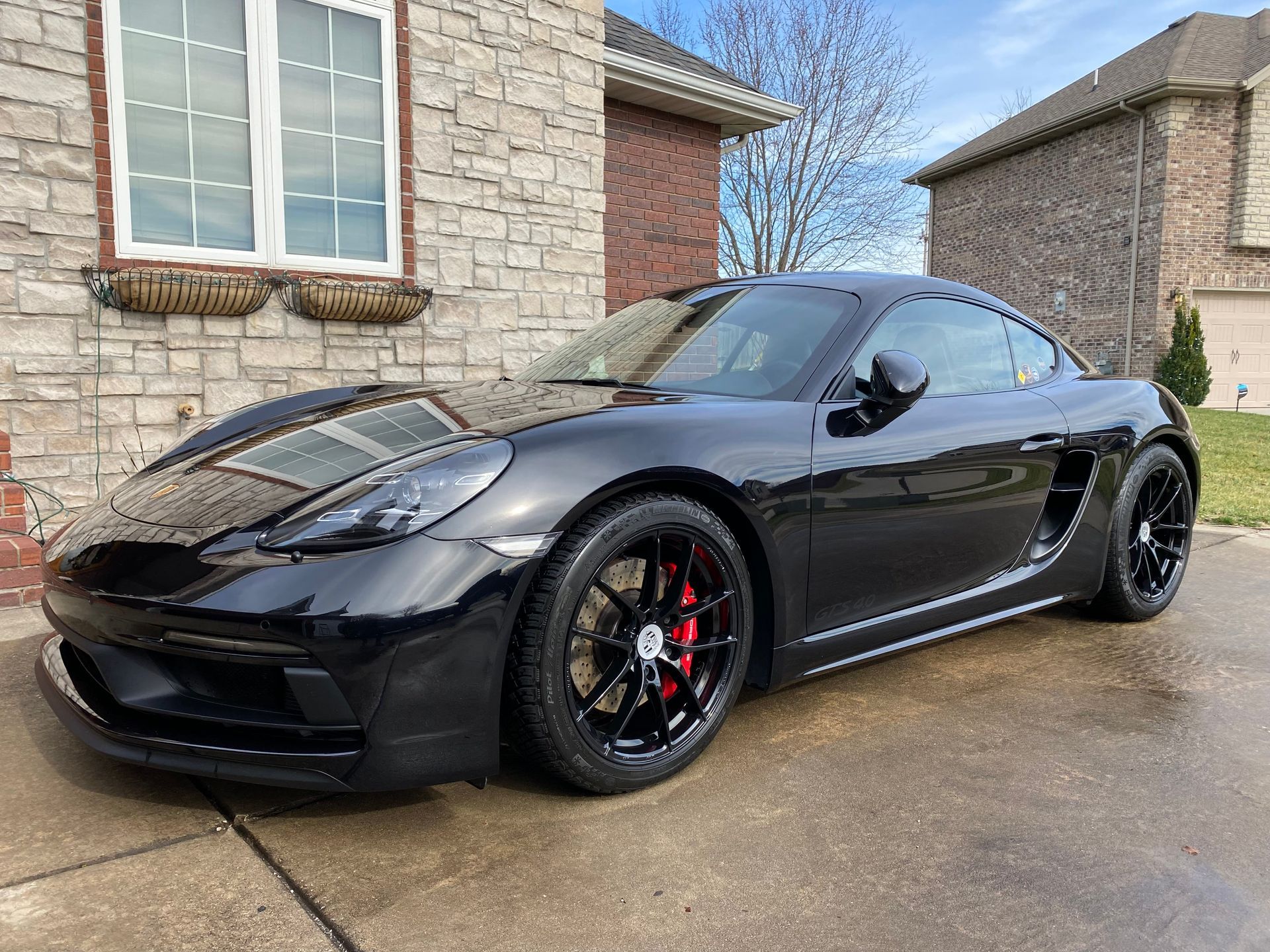 Black sports coupe parked in a driveway beside a stone house, with tinted windows and red brake calipers