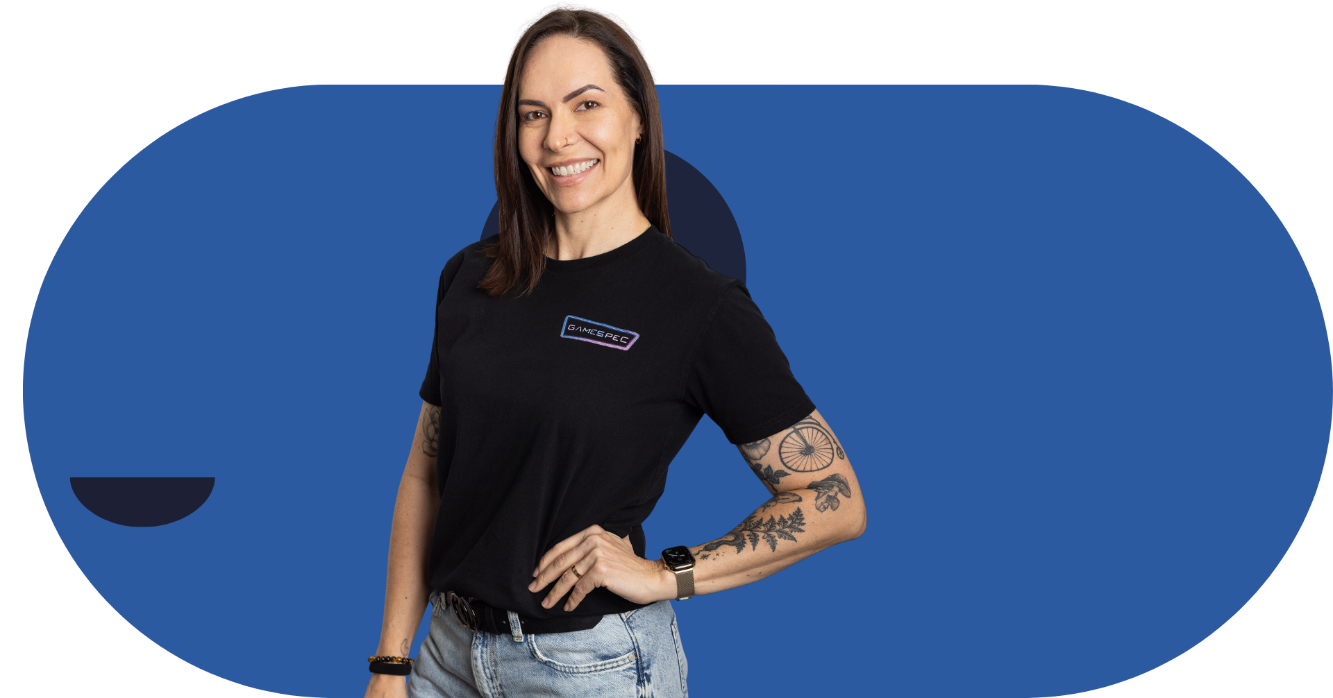 Woman in black T-shirt and jeans standing against a blue background with one hand on her hip
