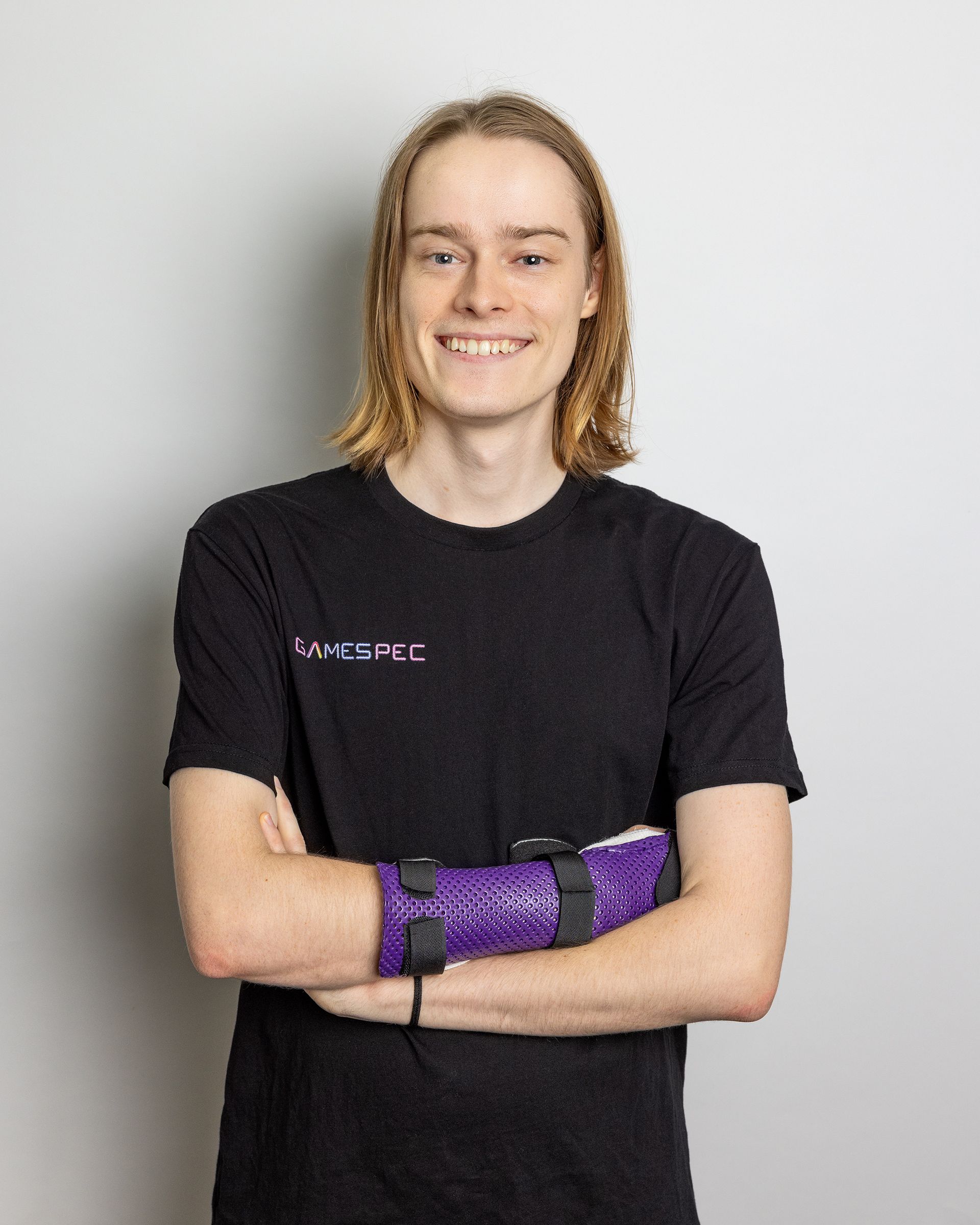 Person standing with arms crossed, wearing a black shirt and purple wrist wraps against a light gray background