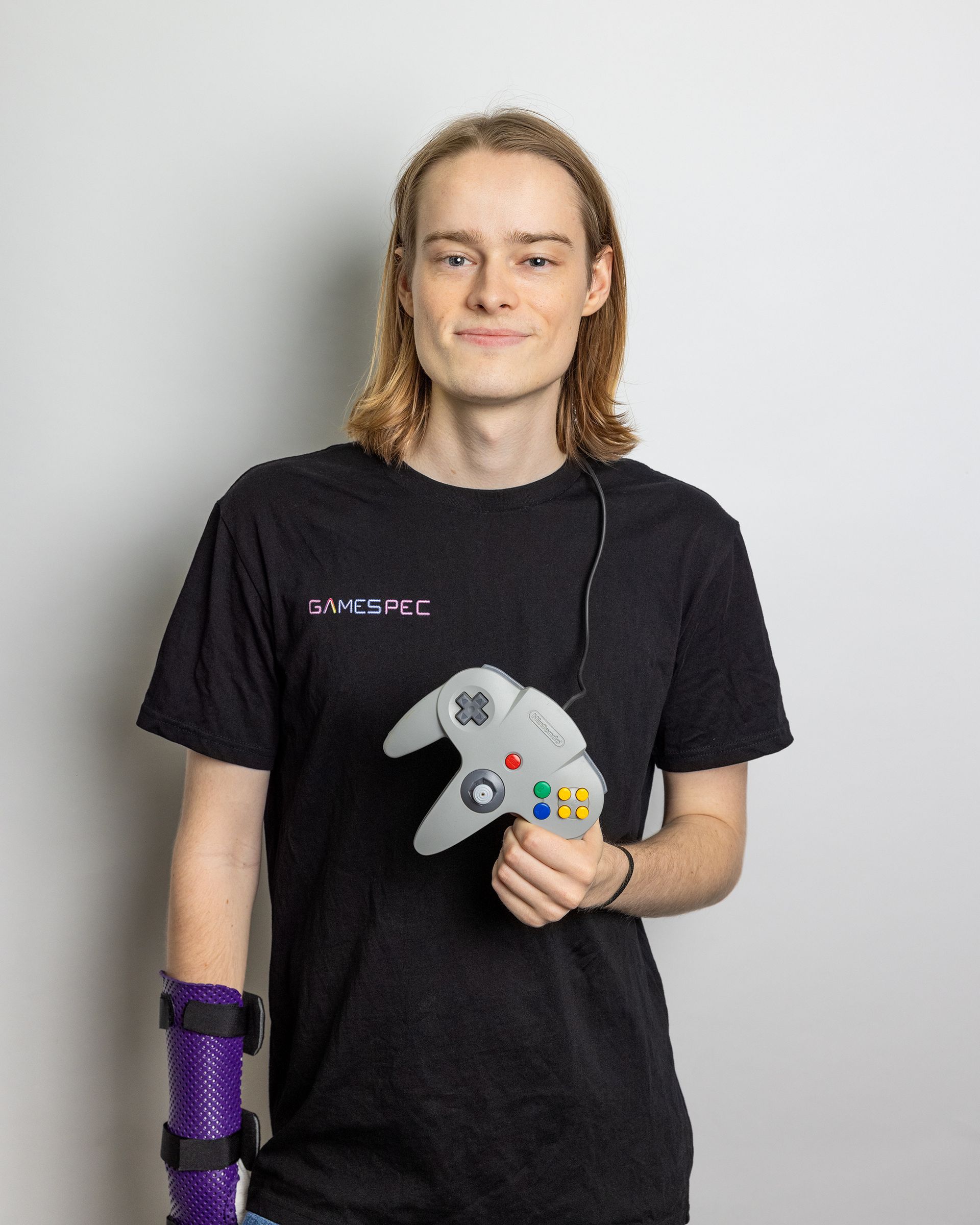 Person in a black T-shirt holding a gray game controller, with a purple brace on one arm, against a plain background.