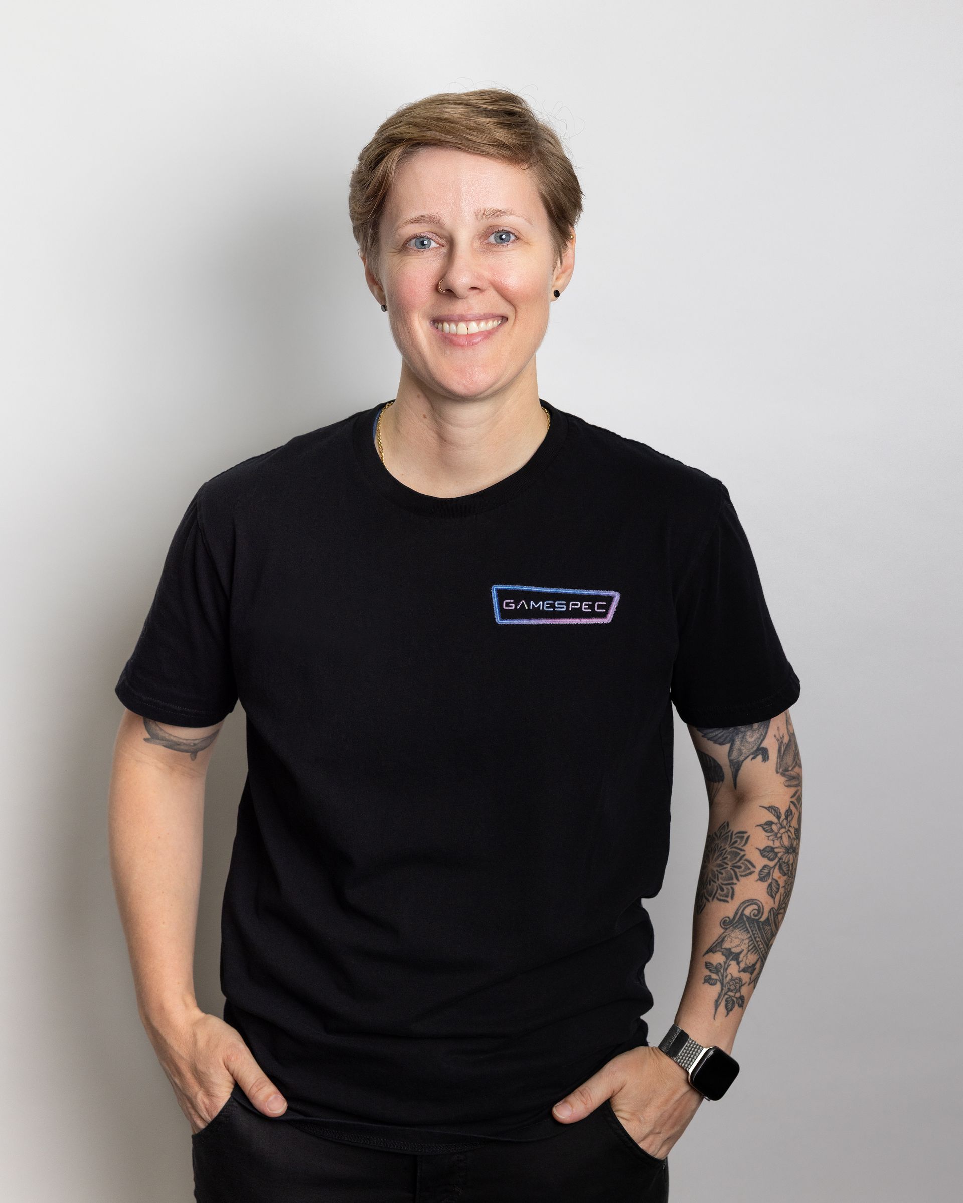 Person in a black T-shirt and pants, smiling with hands in pockets against a light gray background.