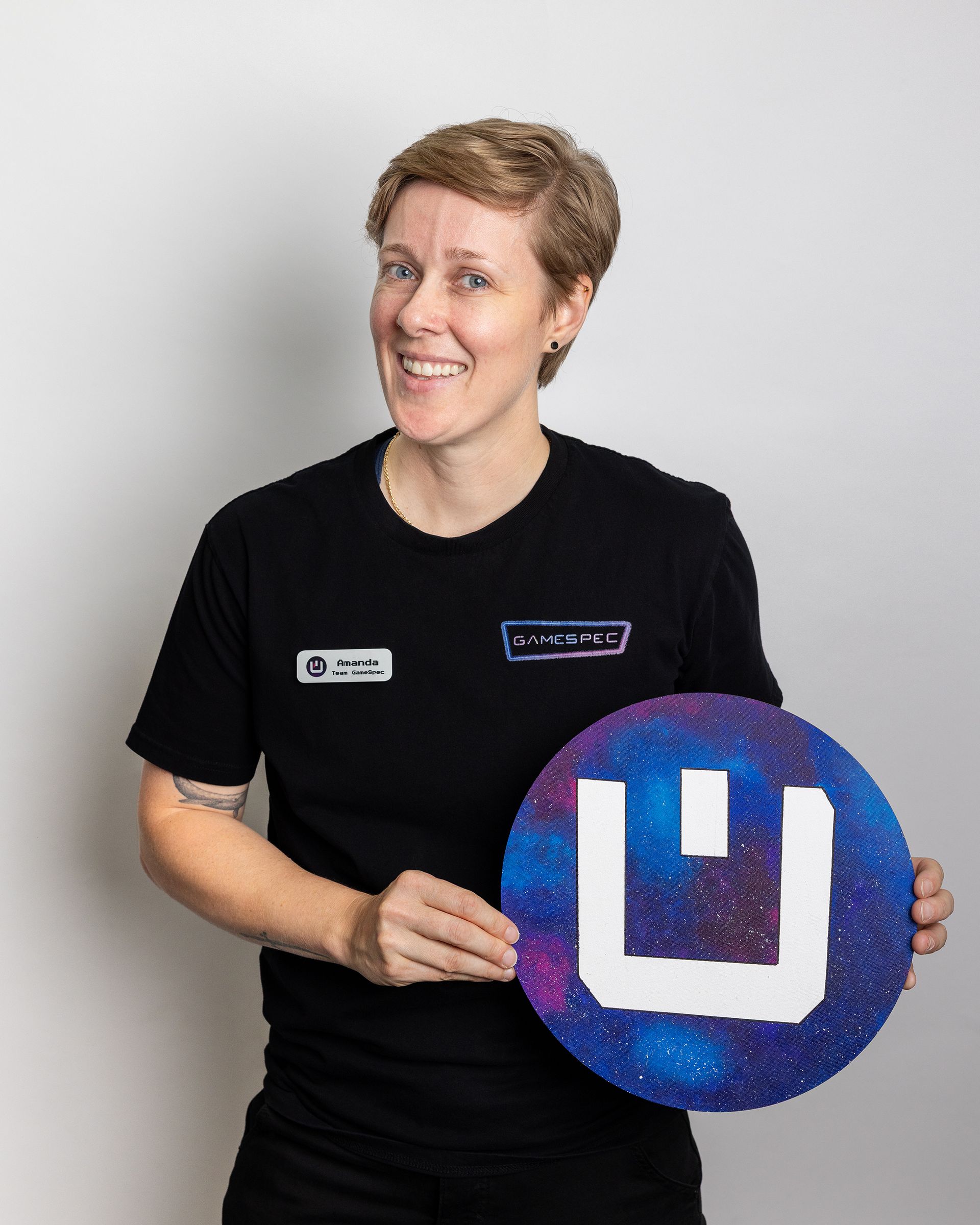 Person in black shirt holding a blue circular sign with a white logo, smiling against a plain background