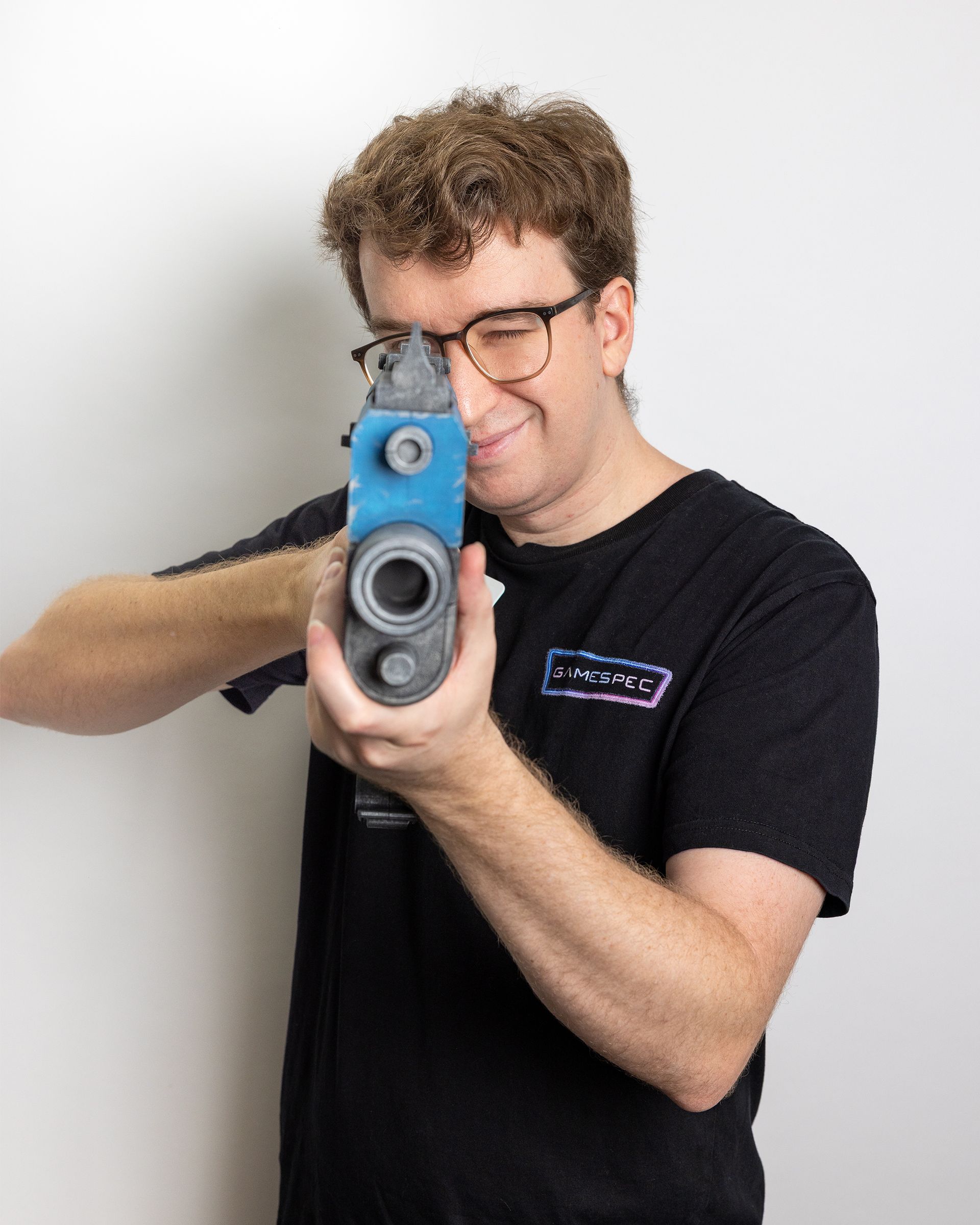 Person aiming a blue toy gun at the camera, smiling, wearing glasses and a black T-shirt.