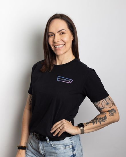 Person smiling in a black T-shirt and jeans, standing against a light gray background with a tattooed arm.