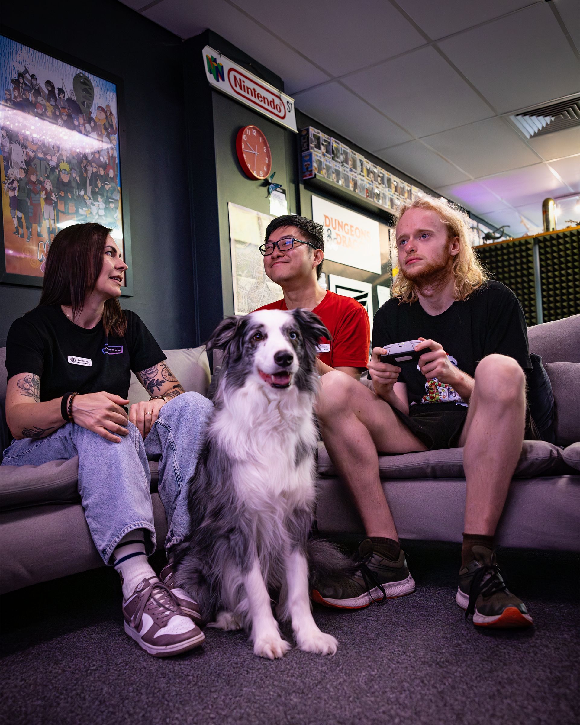 Three people and a white dog sit on a couch in a dim lounge, holding drinks and chatting.