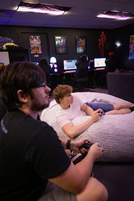 Two people playing video games on a couch in a dimly lit room with multiple monitors.