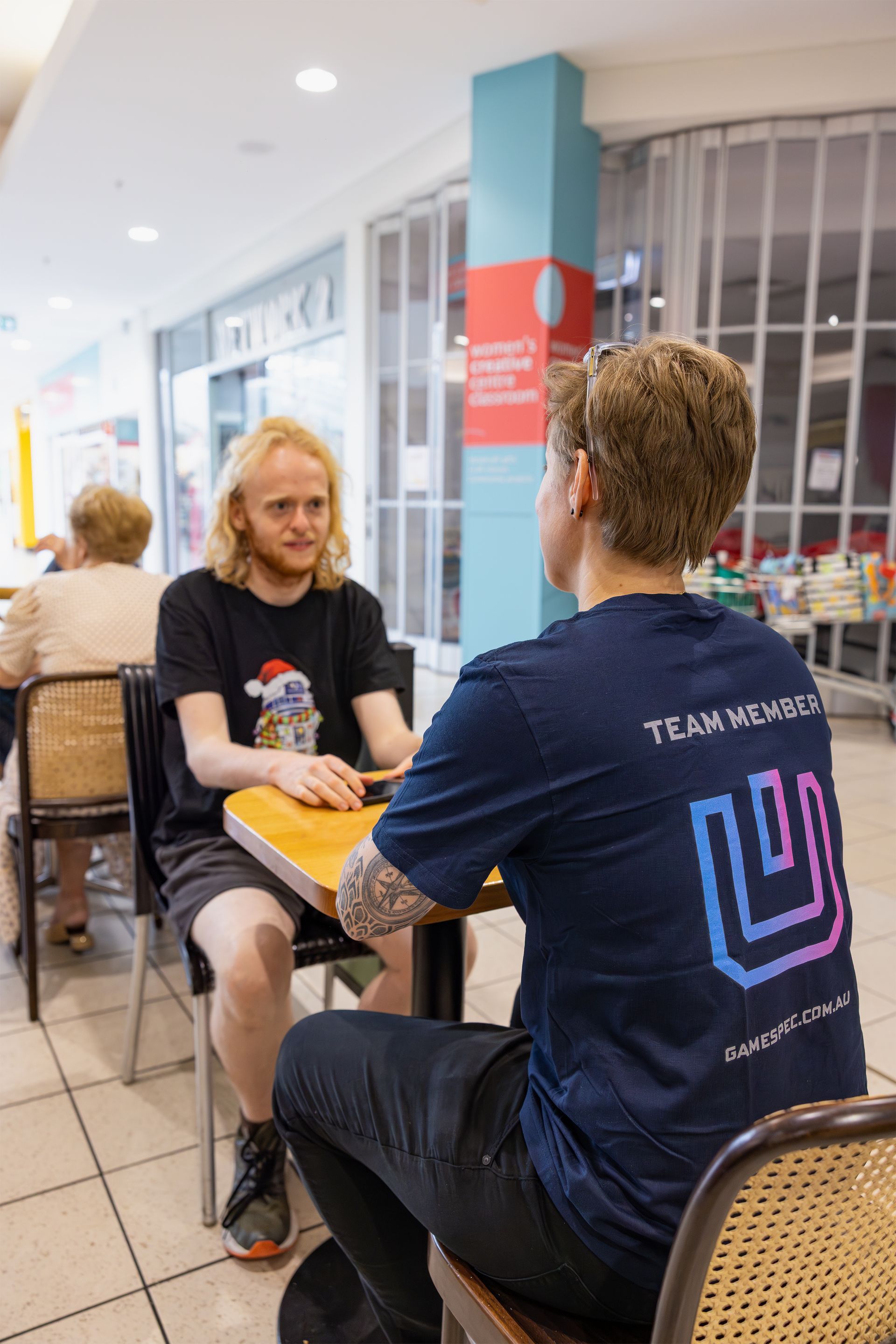 Two people seated at a table in a bright indoor hallway, facing each other in conversation.