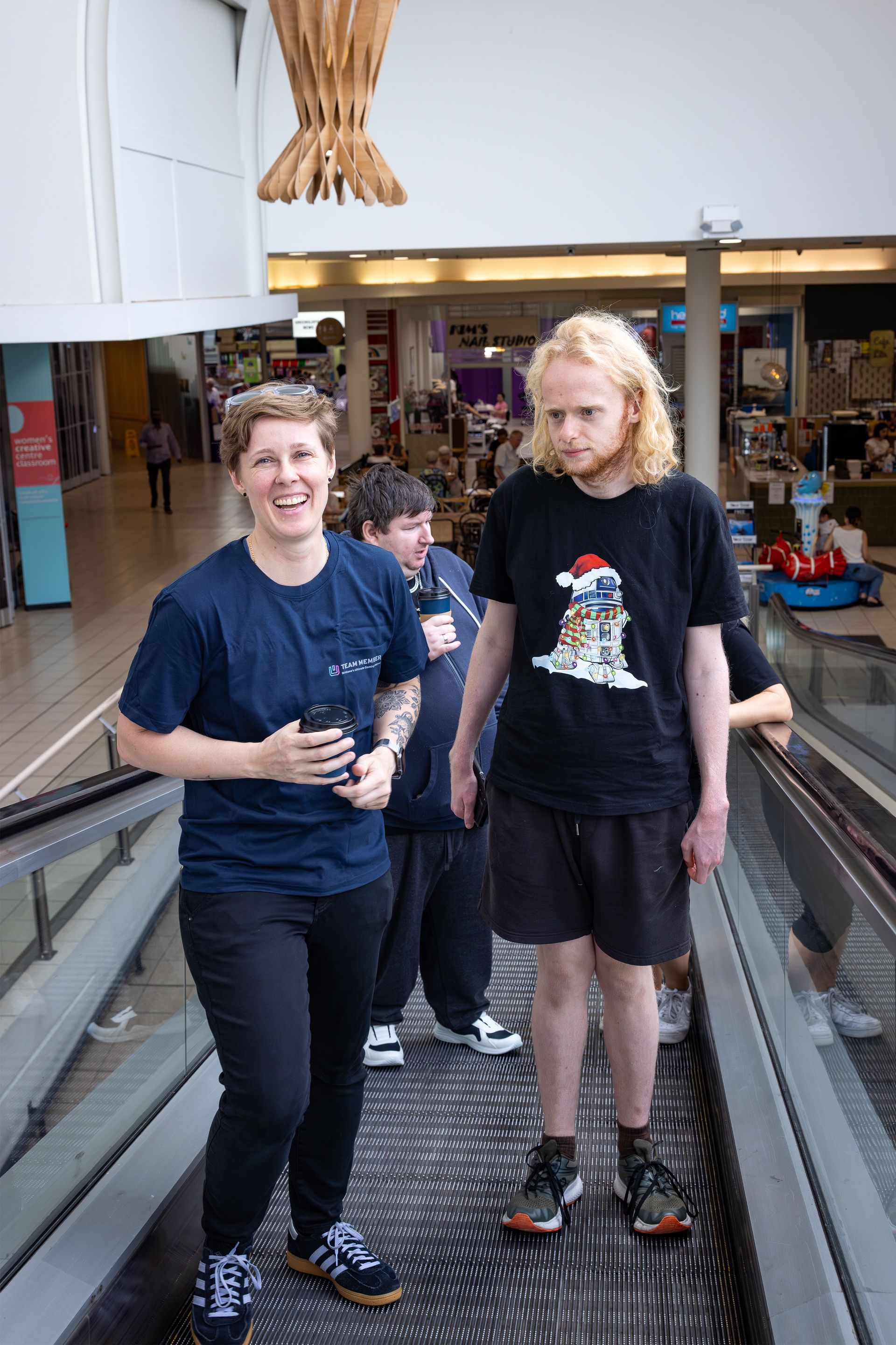 Two people standing on an indoor moving walkway, smiling and looking at the camera.