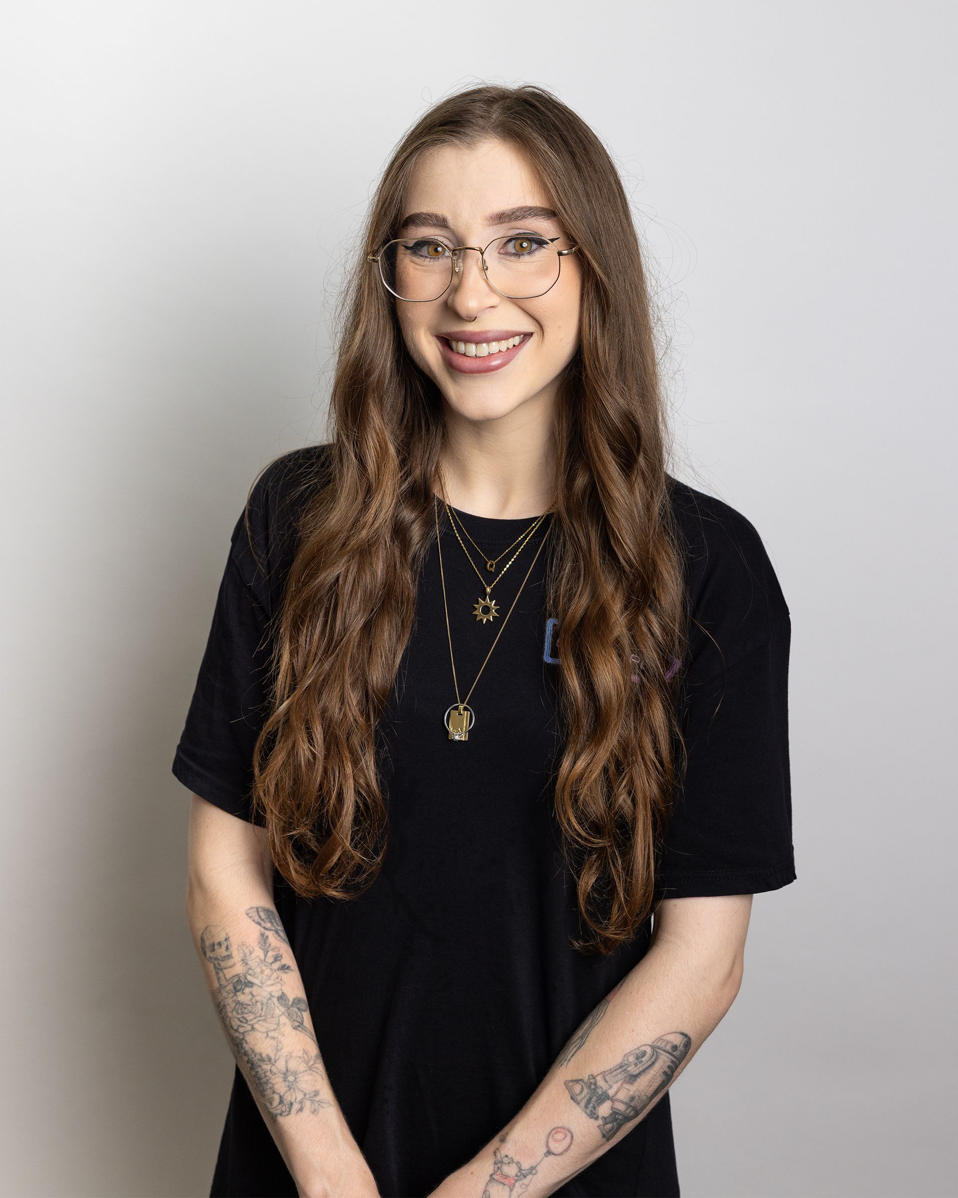 Smiling person with long wavy hair, glasses, black shirt, and tattoos against a light gray background