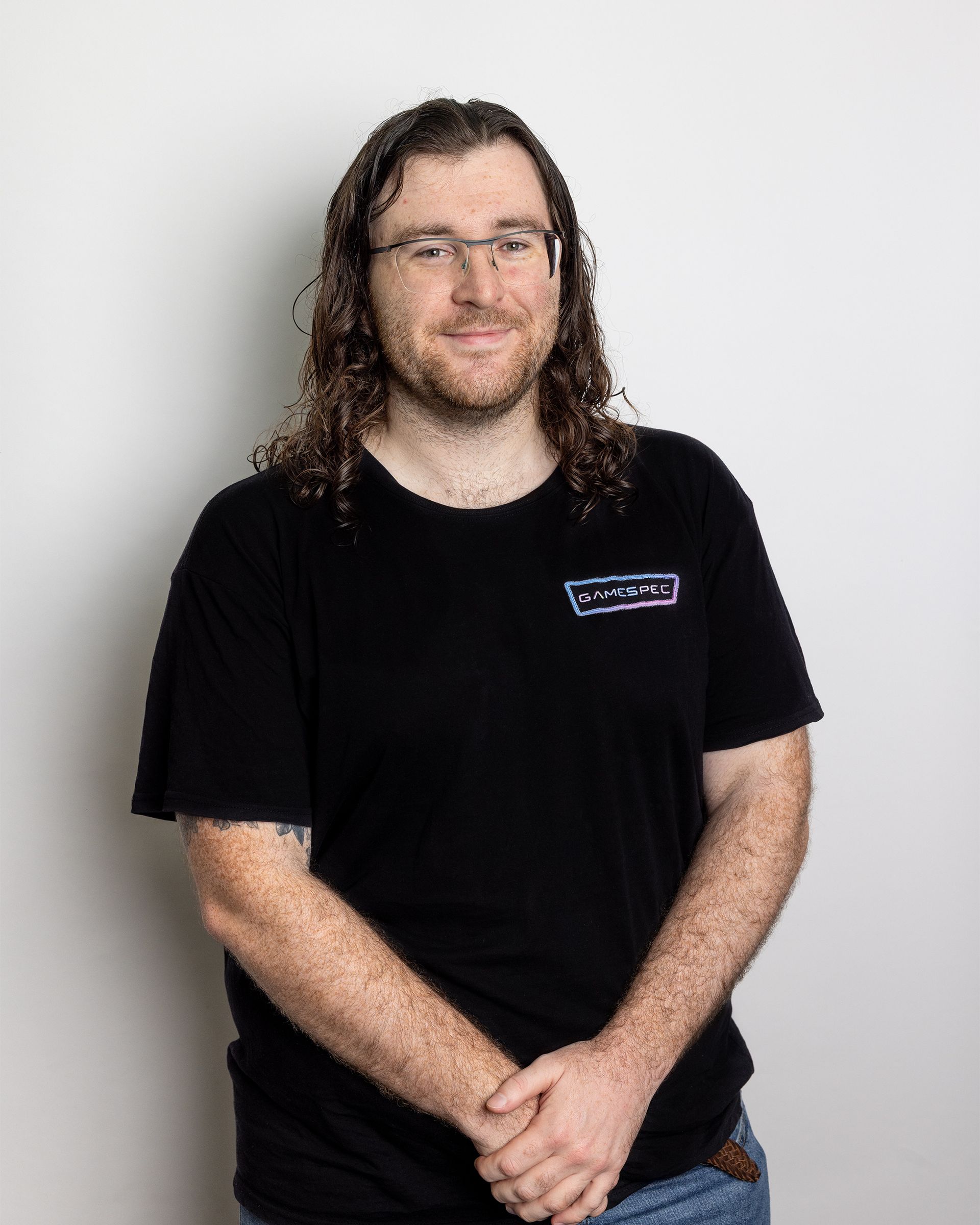 Person with long curly hair and glasses standing against a light gray background, wearing a black T-shirt.