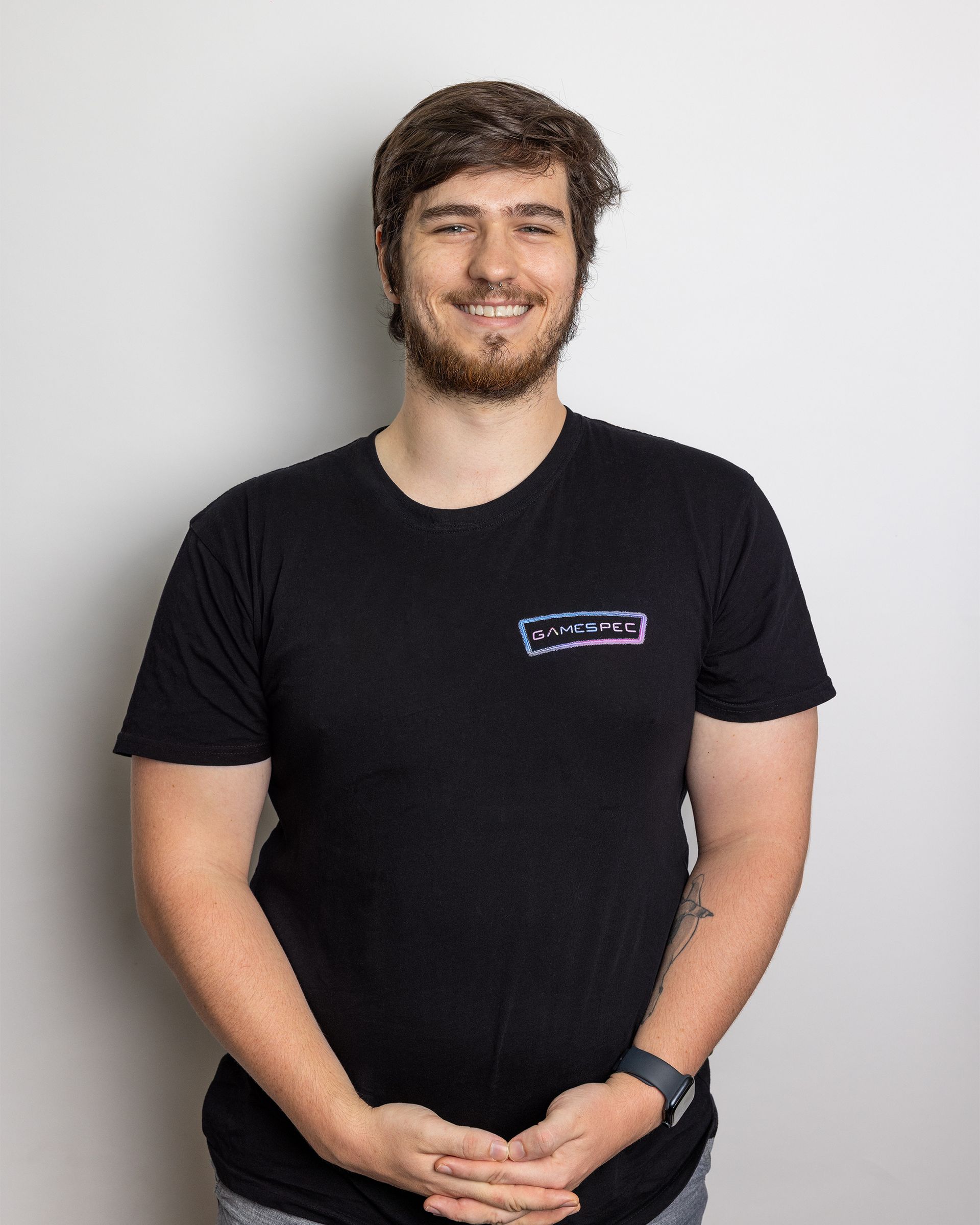 Person standing against a light gray background, wearing a black T-shirt and smiling with hands clasped.