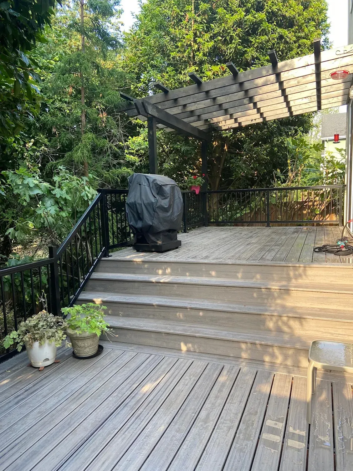 Wooden deck with steps, black grill cover, pergola, and trees in the background.