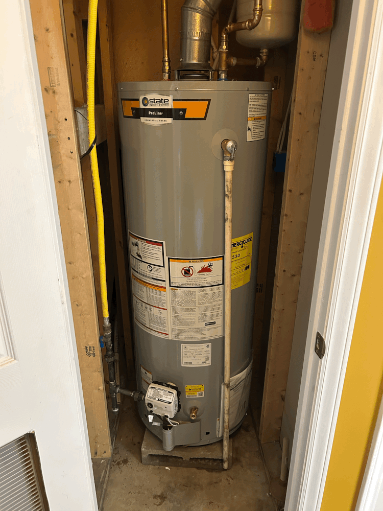 A tall, silver water heater inside a narrow wooden enclosure. Yellow gas line and white drain pipe visible.