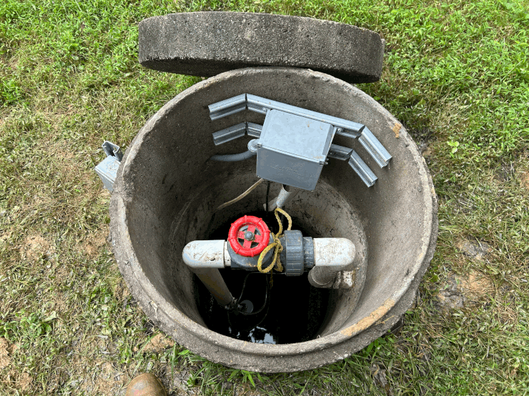 Open concrete well with valve, piping, and electrical components. Lid is partially removed, set in grass.