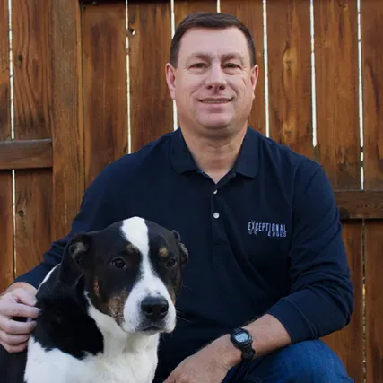 Man seated with black-and-white dog in front of a wooden fence