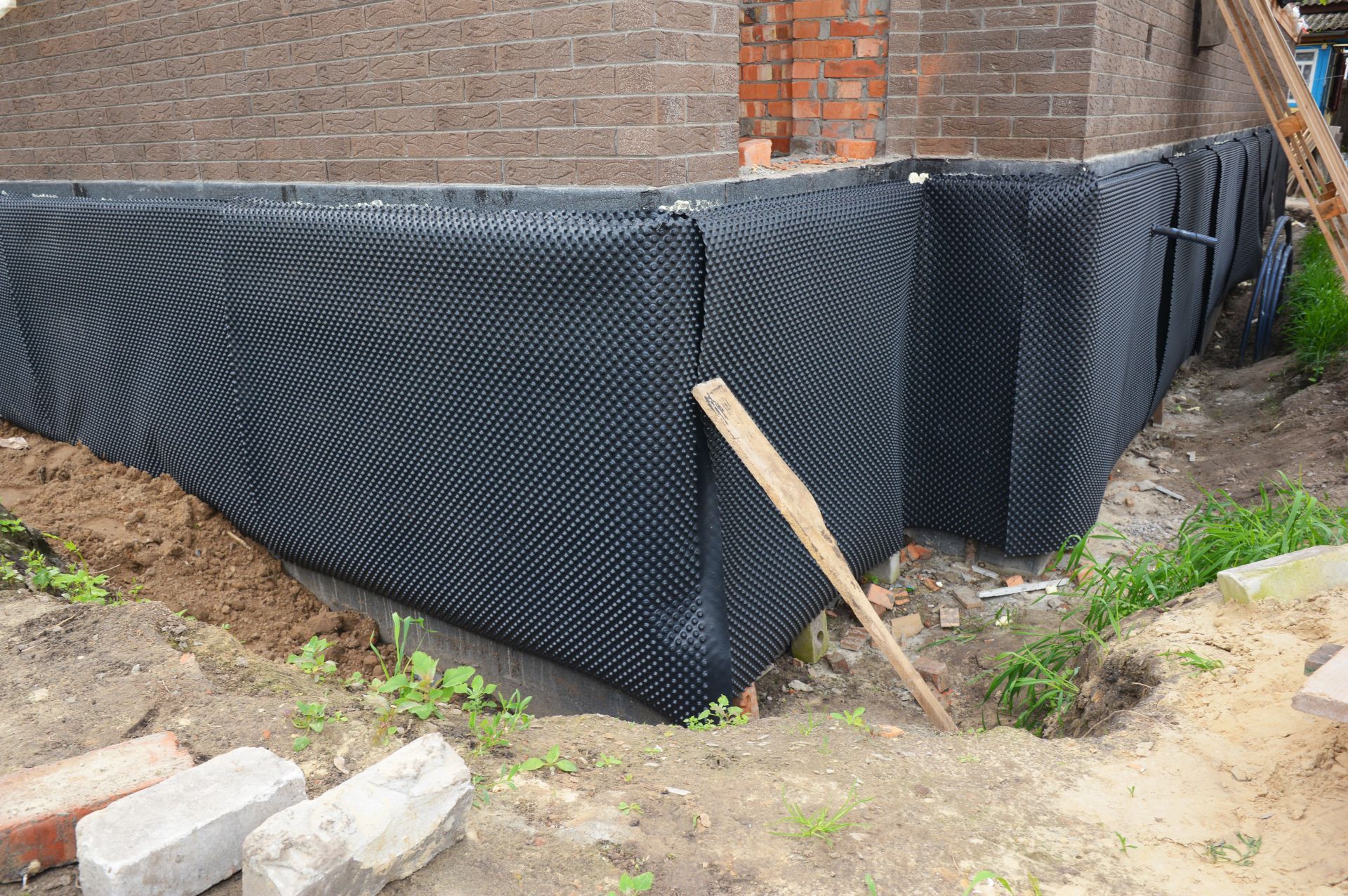 Black foundation wrap around a brick house corner, with a wooden stake and dirt foreground.