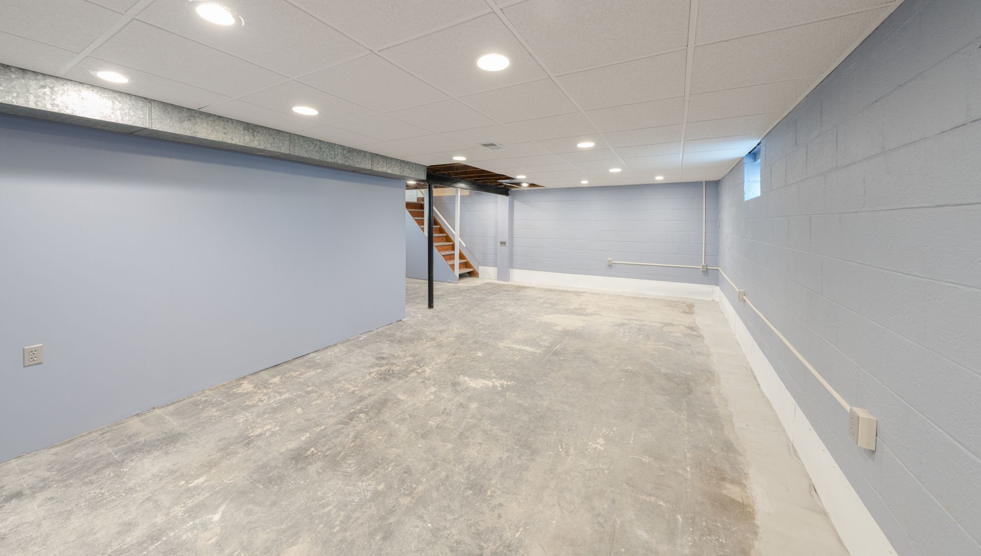 Empty basement room with light blue walls, concrete floor, and stairs at the back