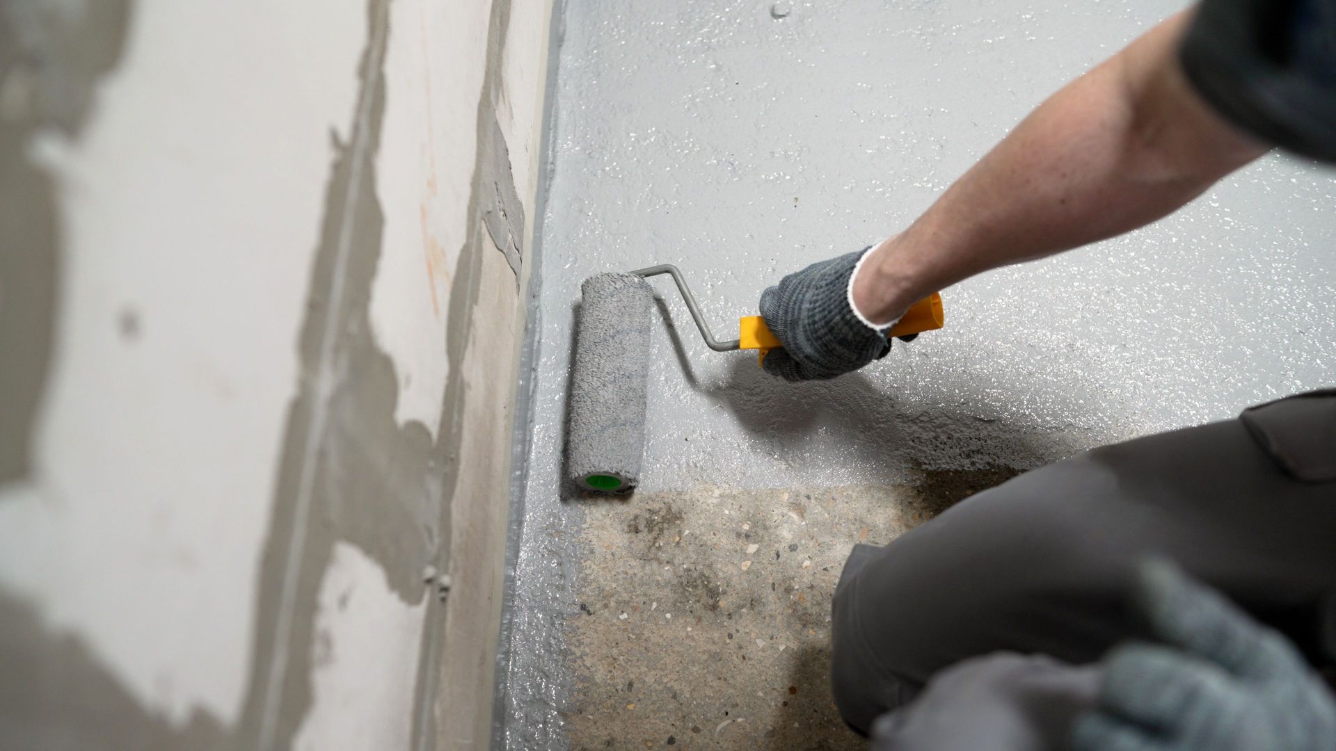 Gloved hand using a trowel to smooth wet concrete along a wall corner indoors