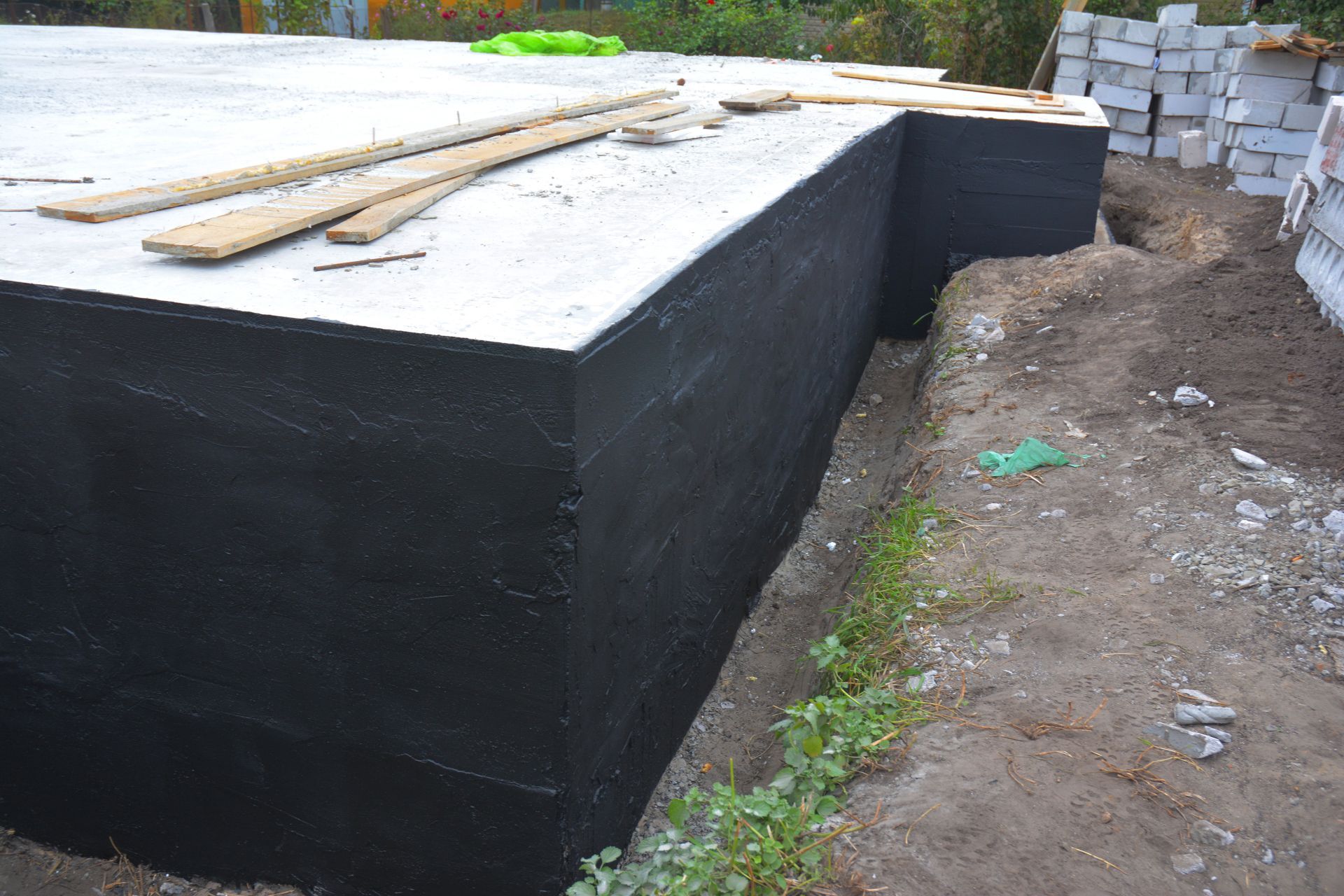 Black waterproofed foundation wall beside a dirt path, with boards on top and stacked blocks in back.