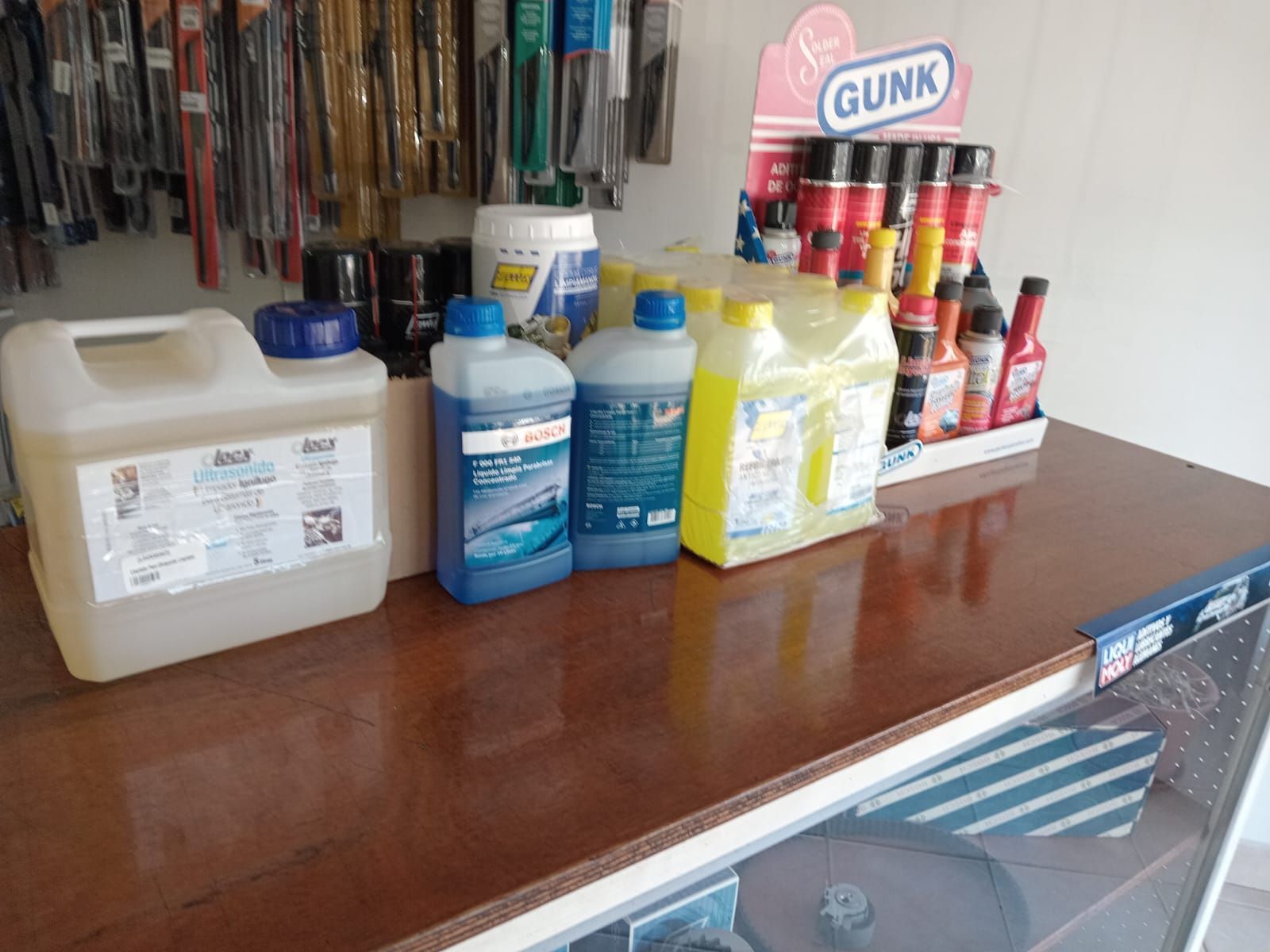 Diversos líquidos y suministros para automóviles sobre un mostrador, con una exhibición de GUNK y limpiaparabrisas en el fondo.