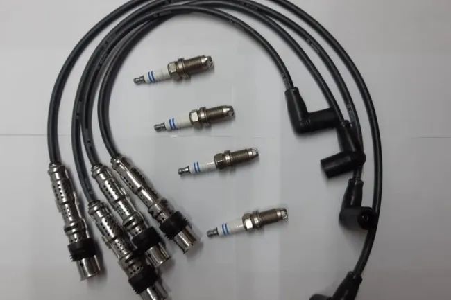 Bujías y cables de bujías negros, probablemente de un motor de automóvil, exhibidos sobre una superficie blanca.
