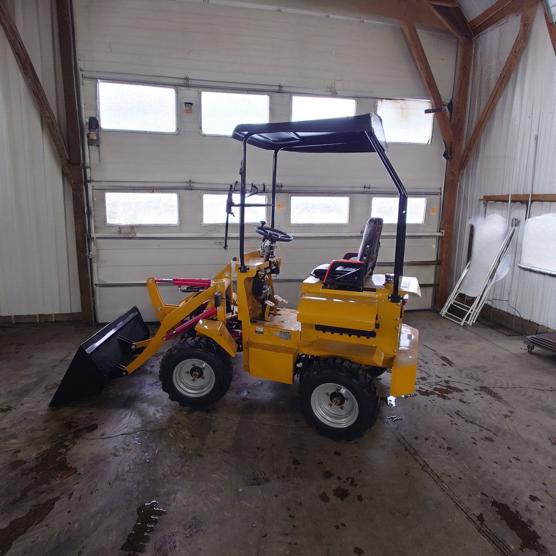 Yellow mini loader with front bucket in a garage, under a white overhead door, with a blue canopy cover