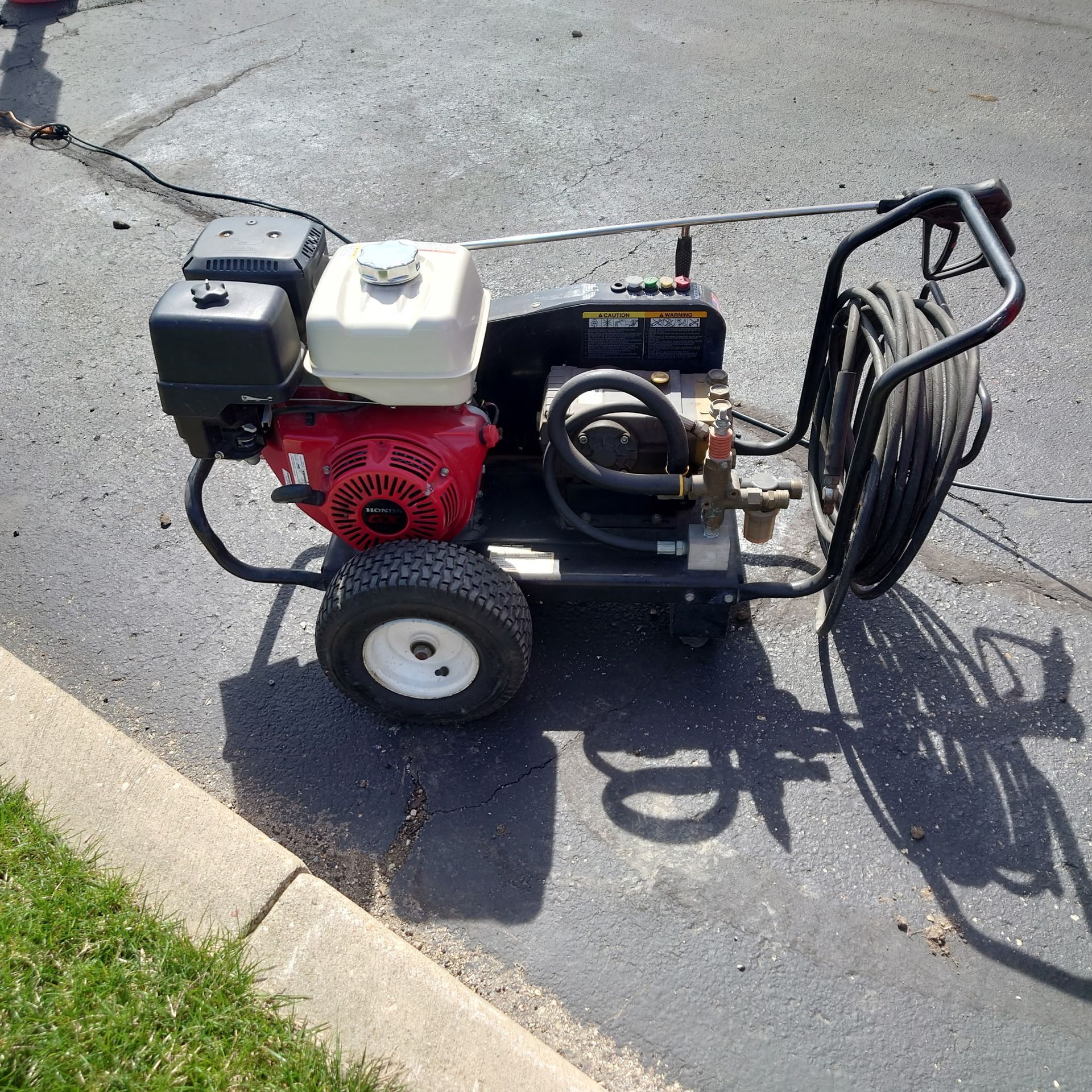 Gas-powered pressure washer on a sidewalk beside a curb, with hose reel and red engine.