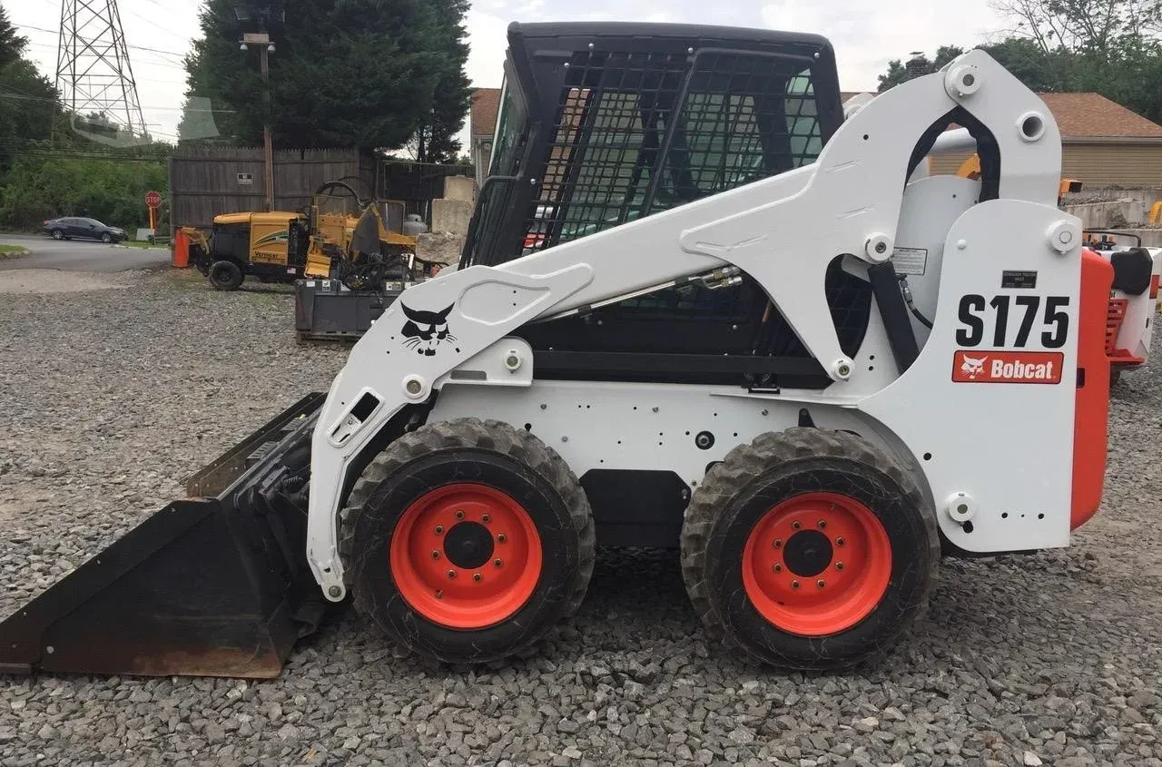 White Bobcat S175 skid steer with front bucket on gravel lot, red wheels, construction equipment in background