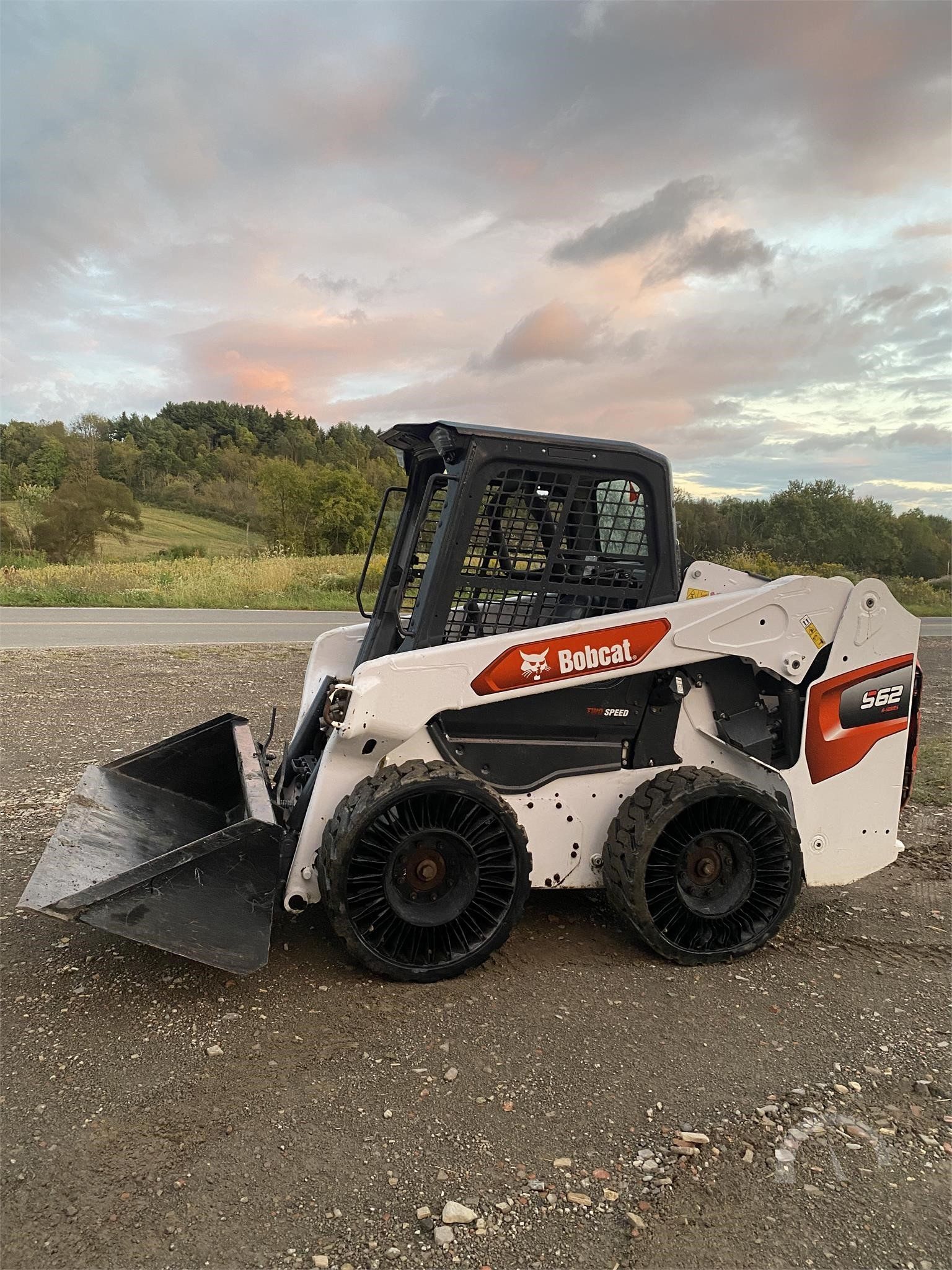 White Bobcat skid-steer loader on gravel road with raised bucket at sunset