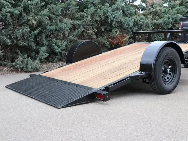 Small black utility trailer with a wooden deck and rear ramp, parked on pavement near bushes