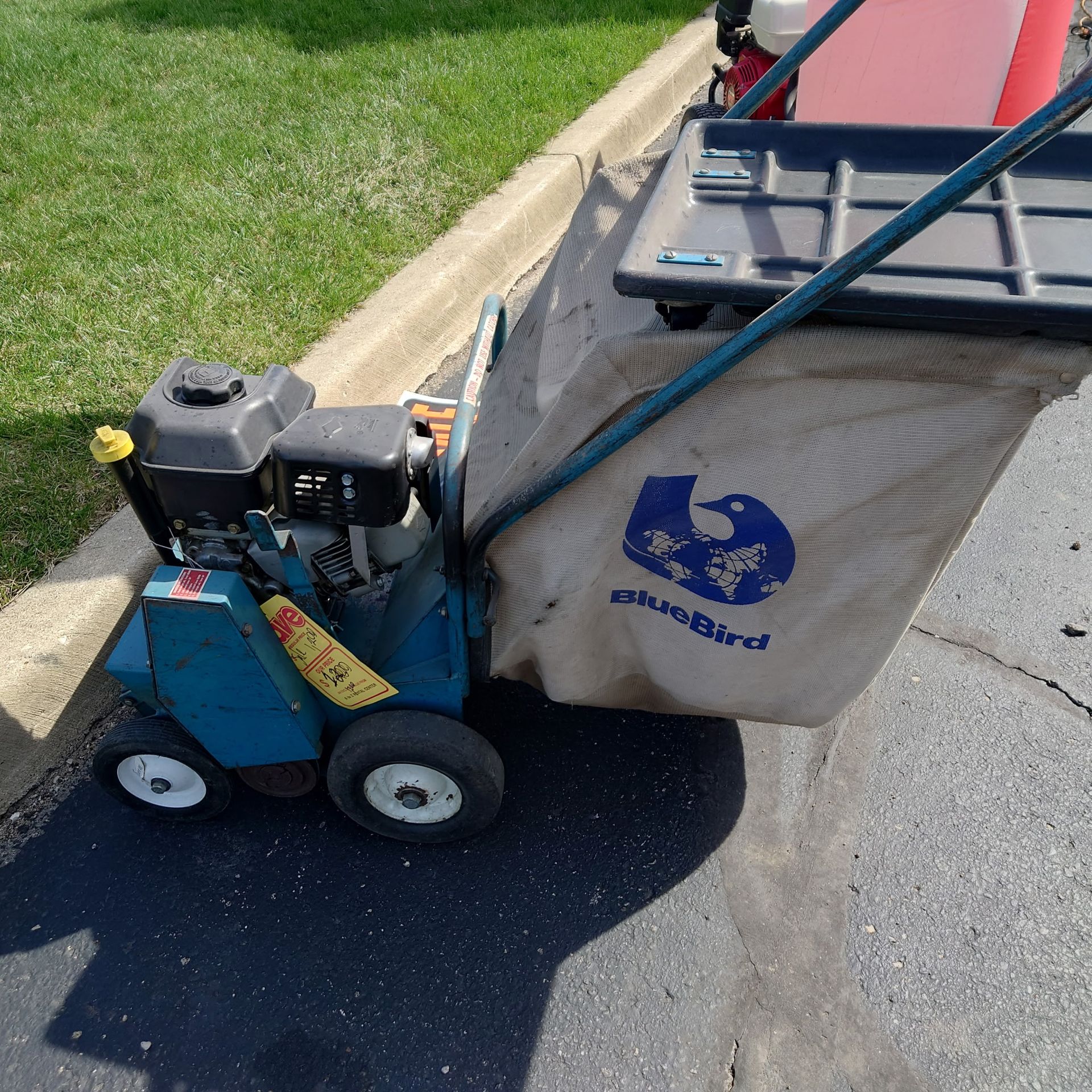 Small blue push lawn mower with a grass catcher bag on a driveway beside a curb and lawn