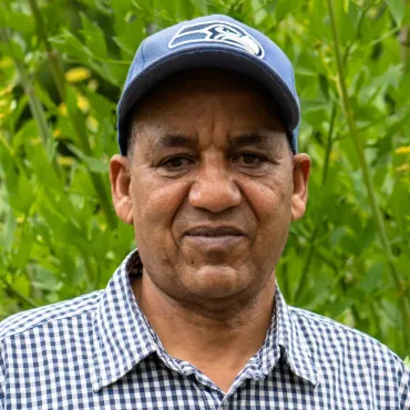 A man wearing a blue hat and a plaid shirt is standing in front of a bush.