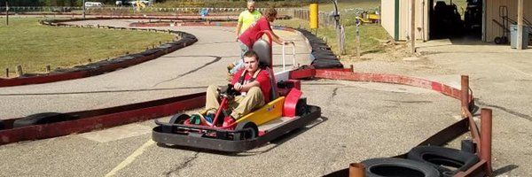 Person in red shirt driving a go-kart on a track, two people watching, and tires nearby.