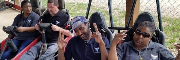 Four people posing for photo inside vehicle, wearing dark shirts with logo. Smiling man in cap gives peace sign. Other woman wears sunglasses.