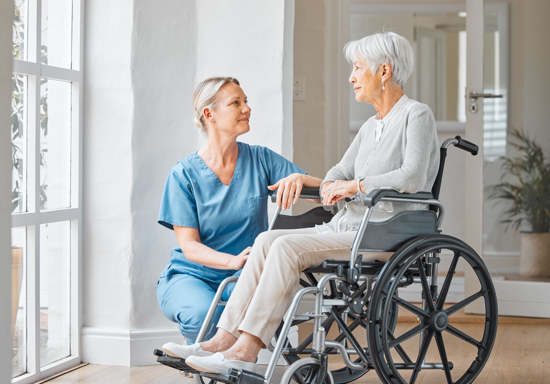 Nurse in blue scrubs talking with a woman seated in a wheelchair near a window.
