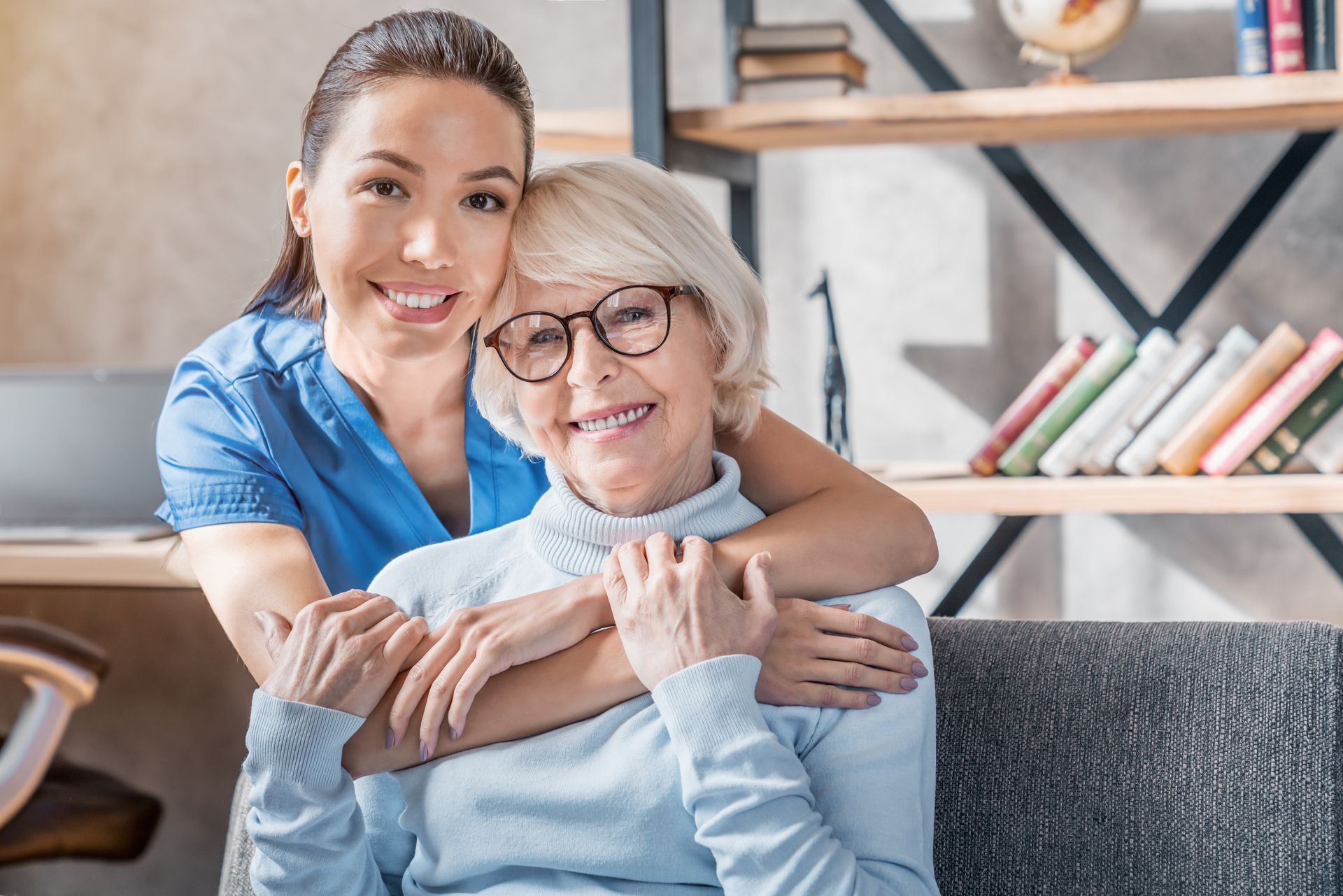 Woman in blue scrubs hugs an older woman wearing glasses; both smile, inside a home.