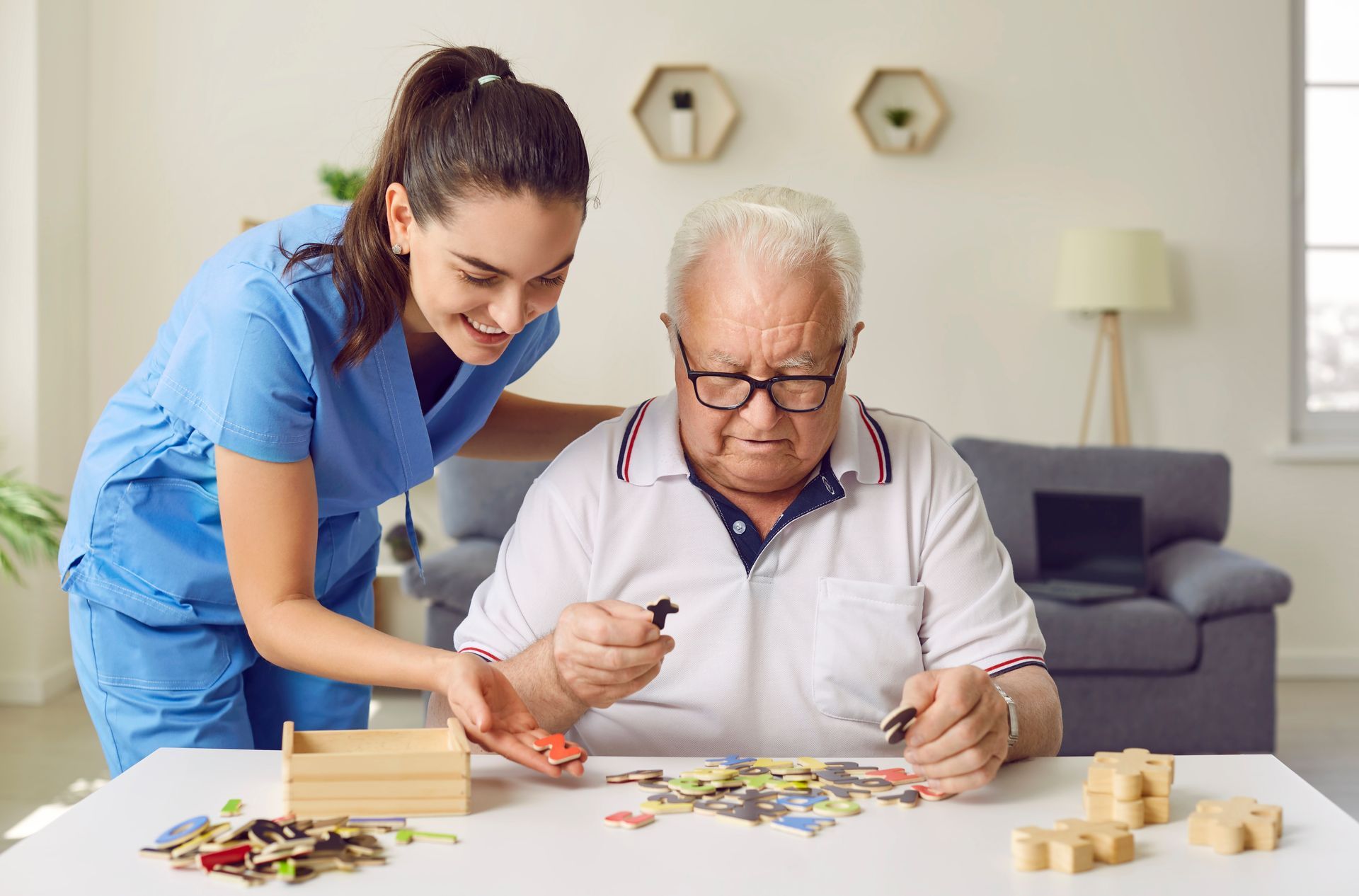 Caregiver assists senior with puzzle. White table, interior setting. Both smiling, caregiver in blue scrubs.