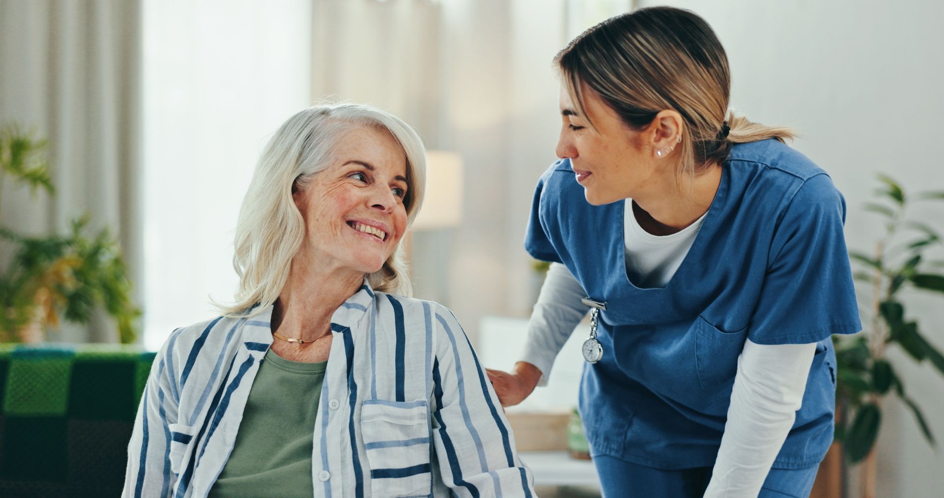 Woman in striped shirt smiles at healthcare worker in blue scrubs indoors.