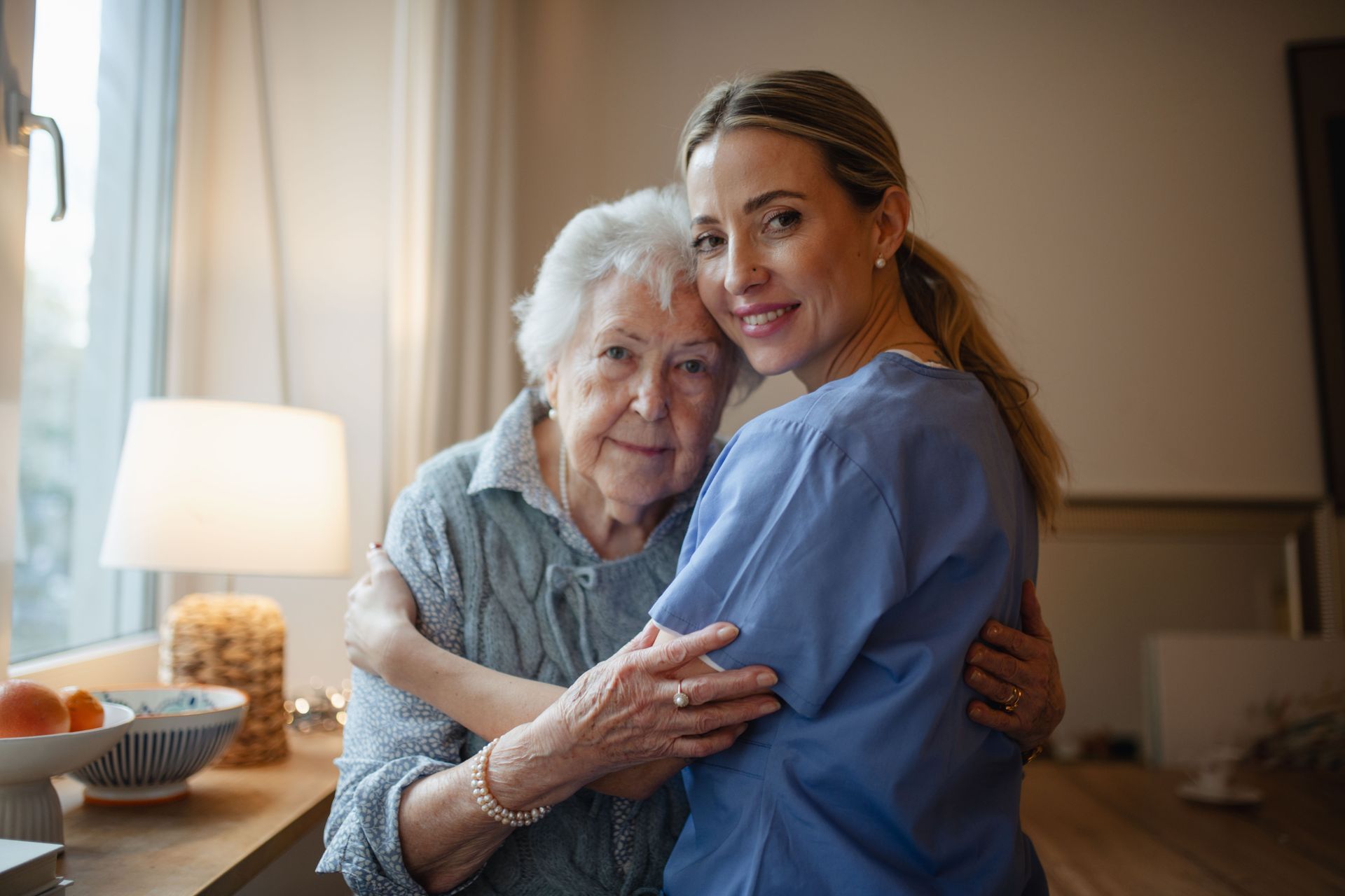 Woman in blue scrubs hugs an older woman; both smile. Indoors, near window and lamp.
