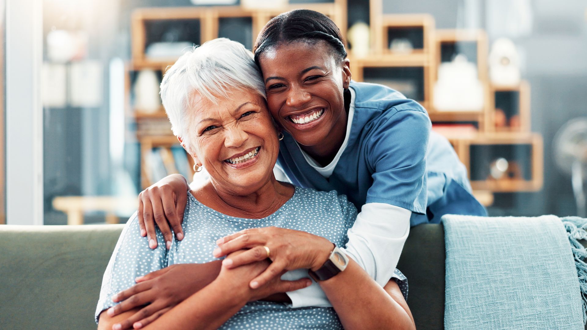 Woman with gray hair embracing a smiling woman in a blue uniform; both indoors.