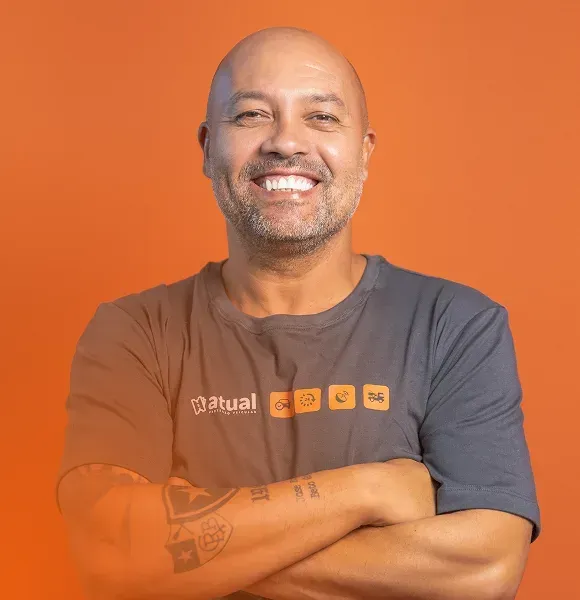 Homem sorridente com os braços cruzados, vestindo uma camiseta cinza escura, contra um fundo laranja.