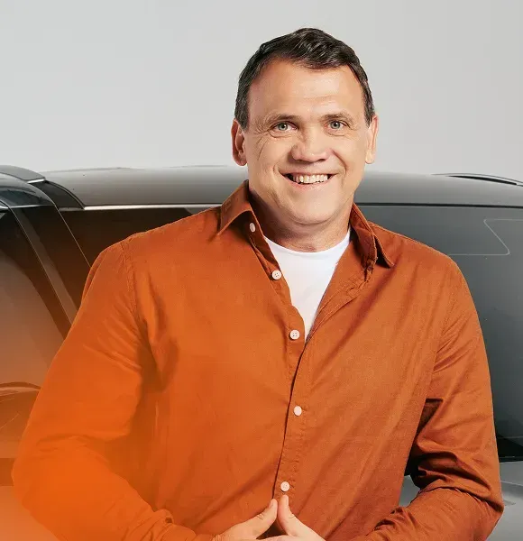 Homem sorrindo, vestindo uma camisa laranja, em pé na frente de um carro preto.