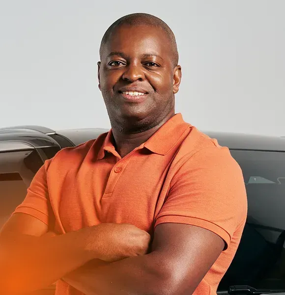 Homem sorridente, vestindo uma camisa polo laranja e com os braços cruzados, está parado ao lado de um carro.