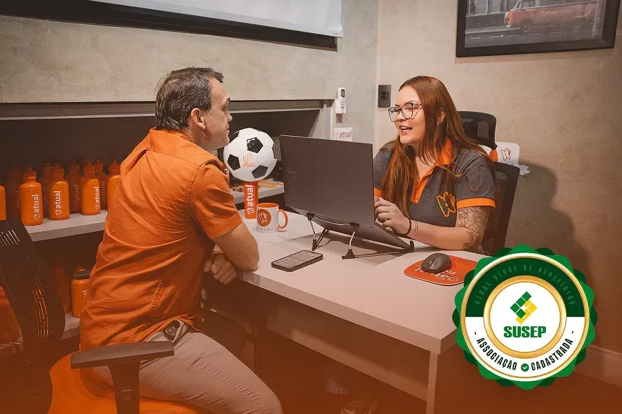 Duas pessoas conversando em frente a uma mesa em um escritório, com um laptop e um troféu de bola de futebol por perto.