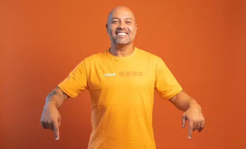 Homem sorridente com camisa laranja aponta para baixo contra um fundo laranja.