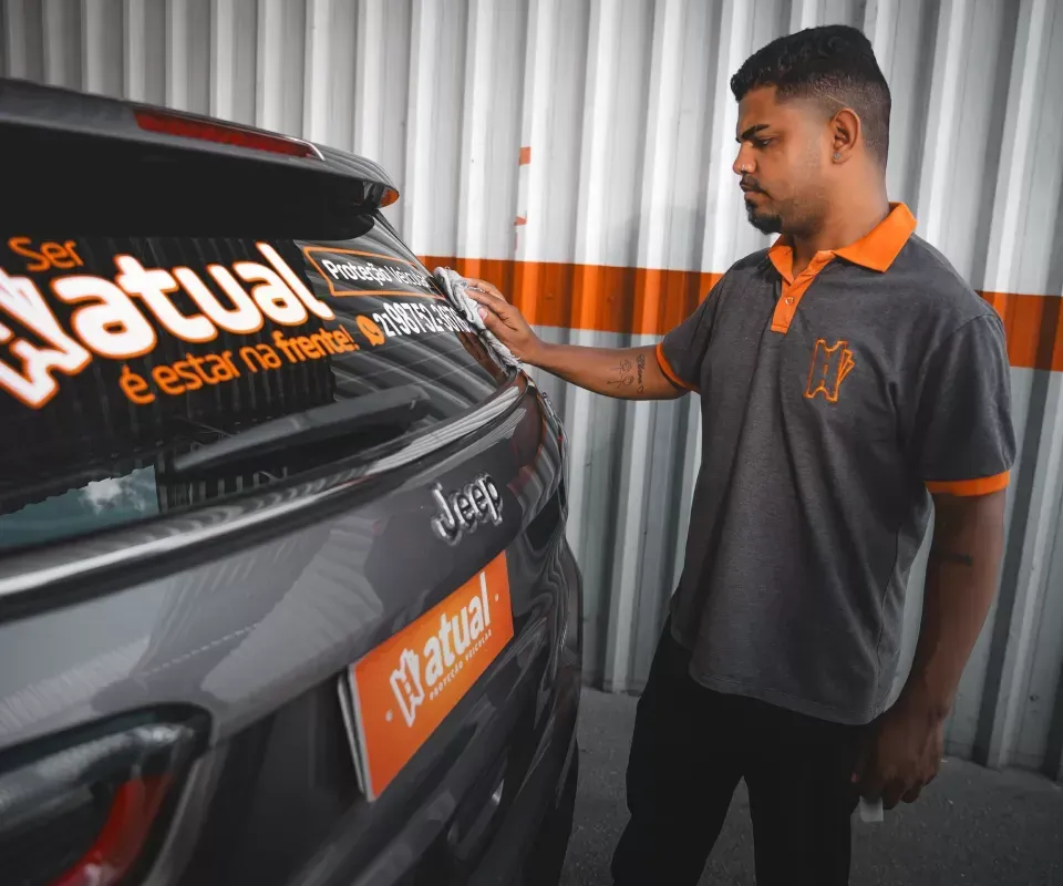 Homem de uniforme cinza e laranja inspecionando um carro preto com detalhes em laranja em uma garagem.